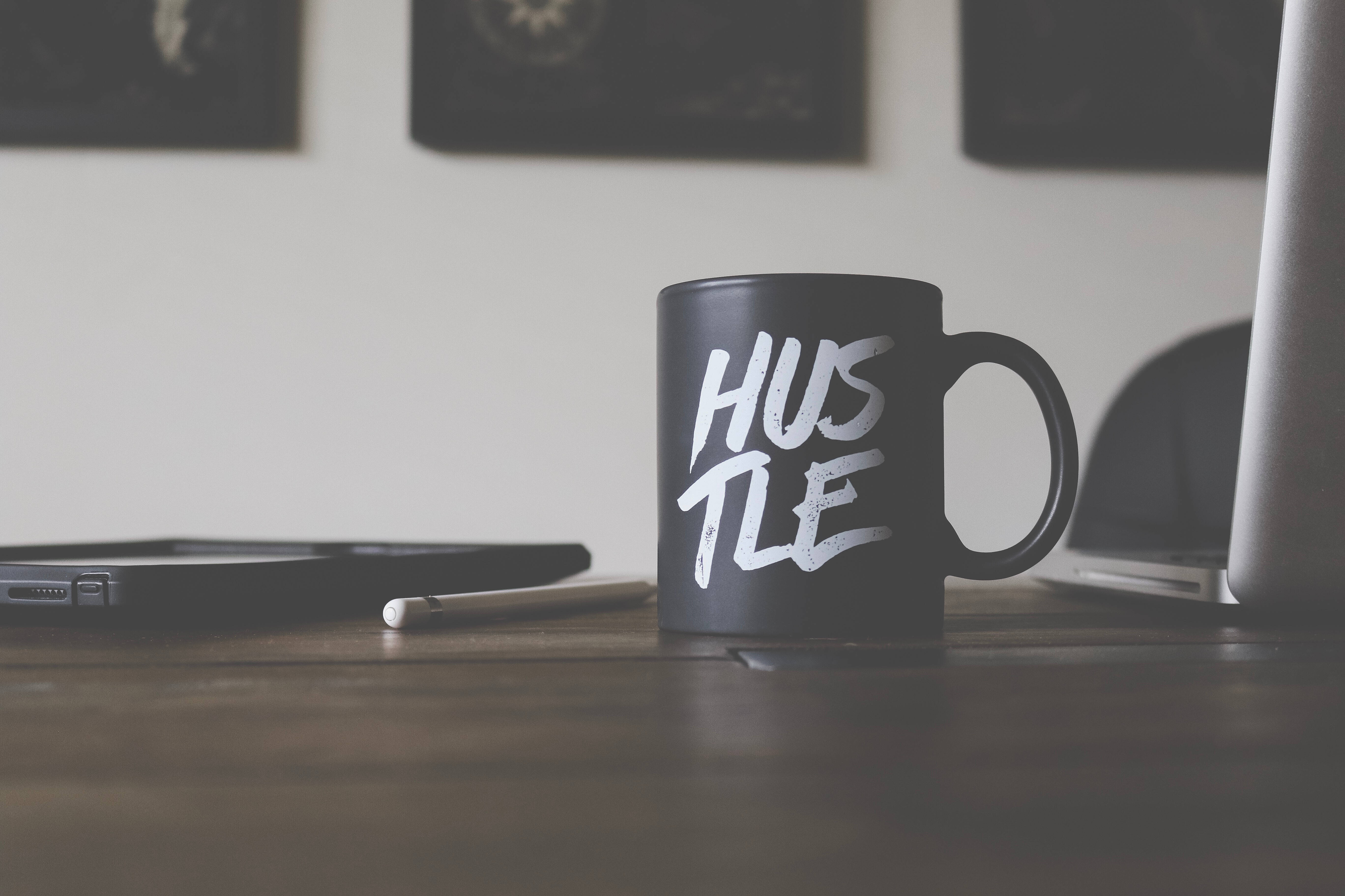 平板電腦 桌布 壁紙 — 一個黑色馬克杯帶有白色 'HUSTLE' 文字，放在木桌上，旁邊有筆記型電腦和平板電腦，營造簡約工作空間。