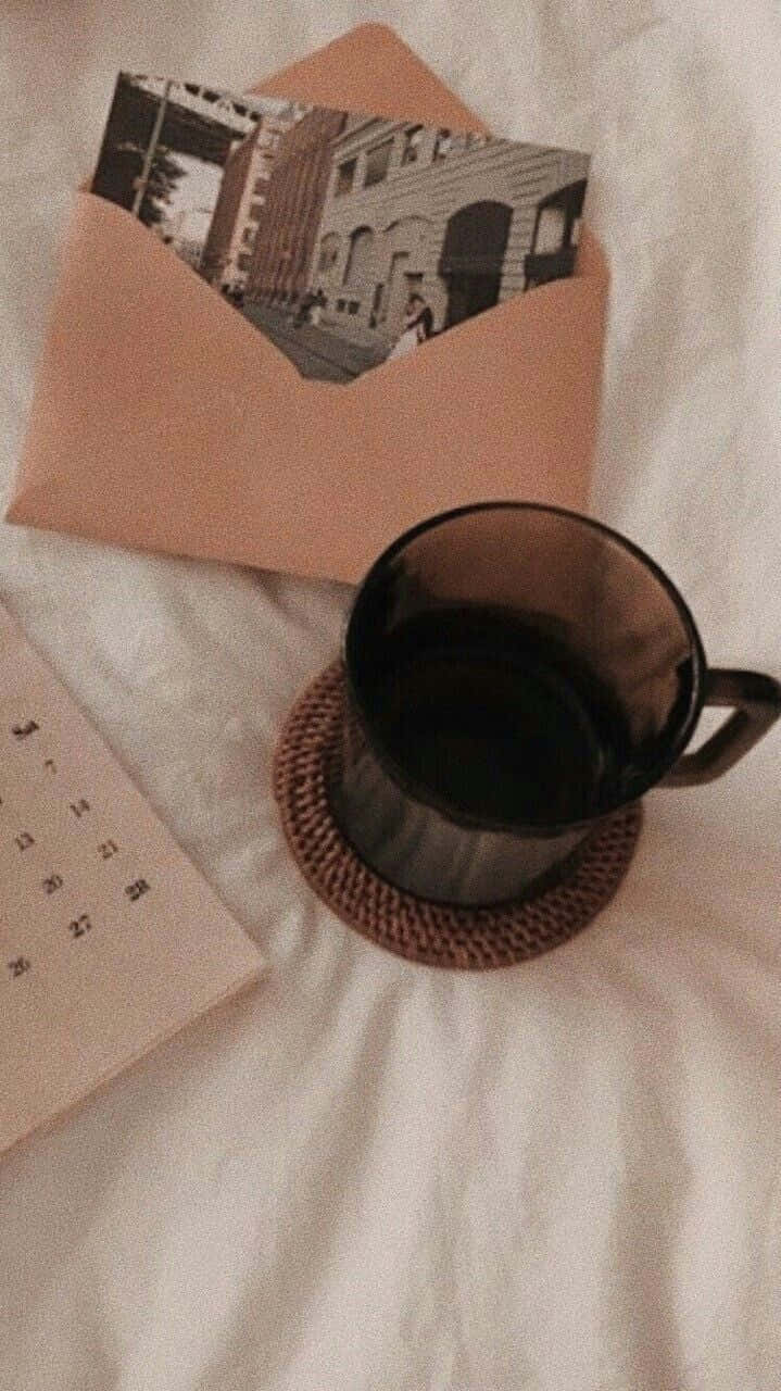 淺棕色美學動圖 桌布 壁紙 — 一個黑色馬克杯放在編織杯墊上，旁邊有一個桃色信封，內含一張復古街頭照片和一本筆記本，置於柔軟的白色布料上。