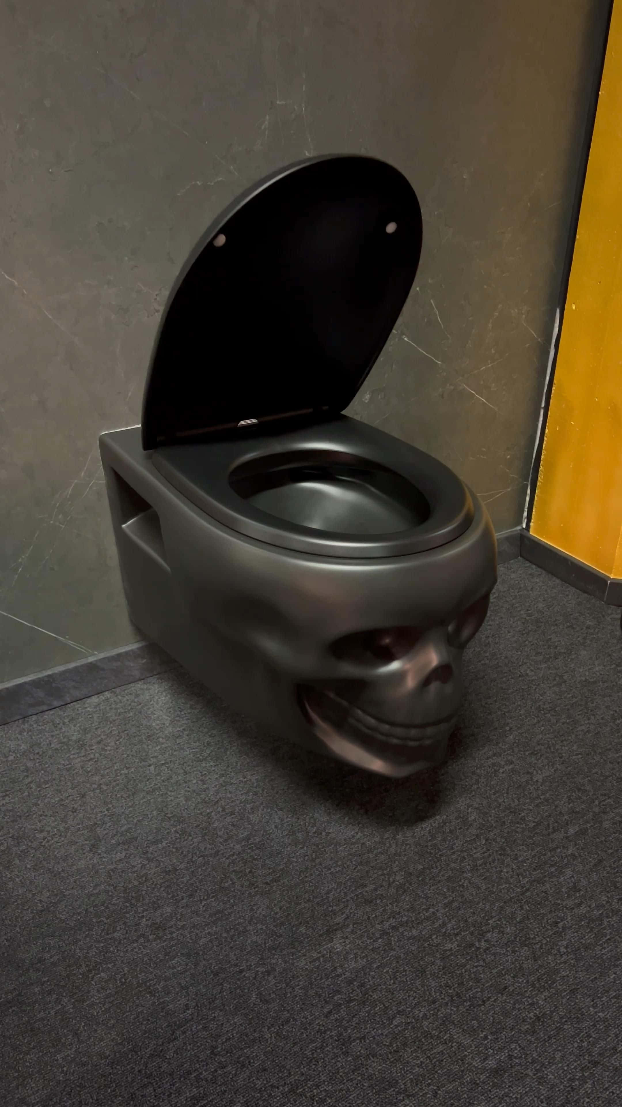 Toilet 桌布 壁紙 — 一個黑色骷髏形狀的馬桶，打開蓋子，放在光線昏暗的浴室角落，背景是灰色牆壁和地毯。