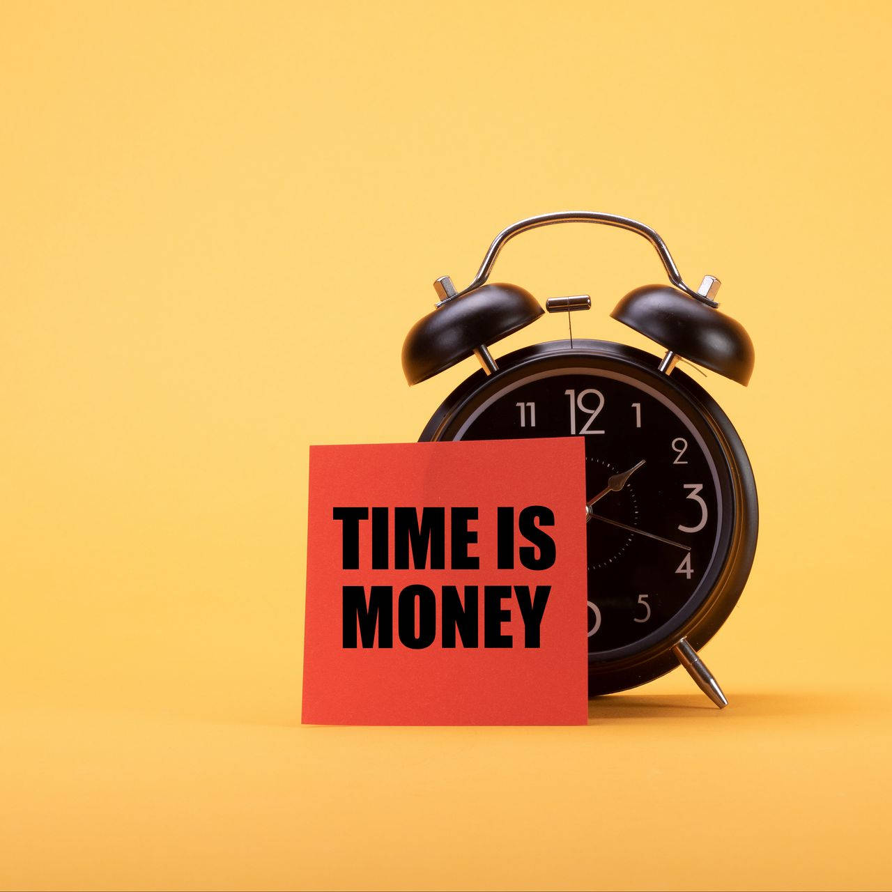 Alarm 桌布 壁紙 — 一個黑色鬧鐘站在一張紅色卡片旁邊，卡片上寫著 time is money，背景為純黃色。