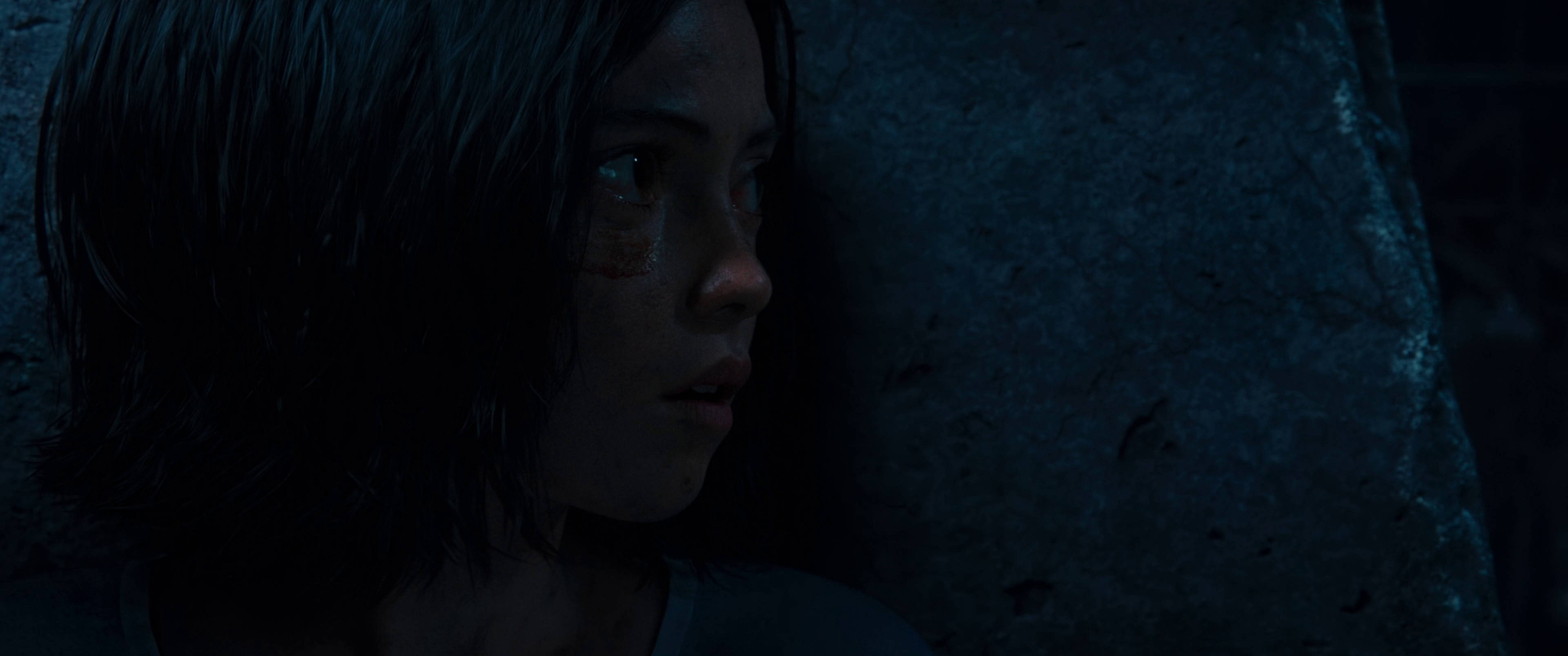 Alita Battle Angel 桌布 壁紙 — 一個黑髮、臉頰有瘀傷的年輕女孩，在昏暗的藍光中靠著粗糙的石牆。