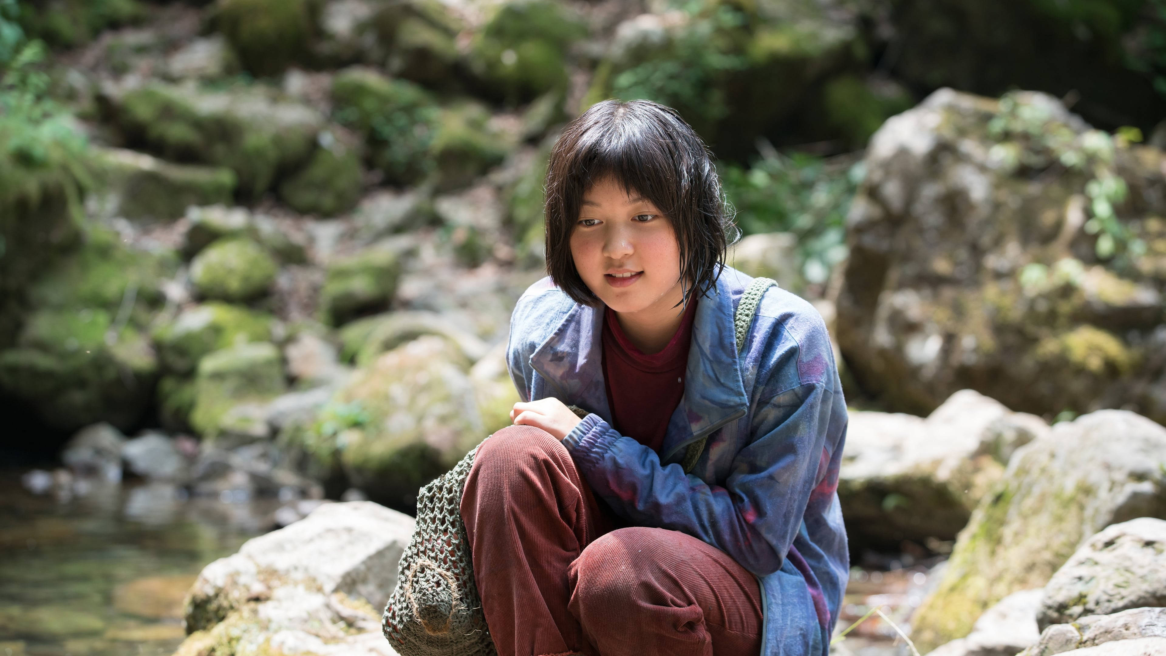 okja 桌布 壁紙 — 一個黑髮女孩盤腿坐在陽光普照的森林中清澈溪流旁的長滿苔蘚的石頭上。