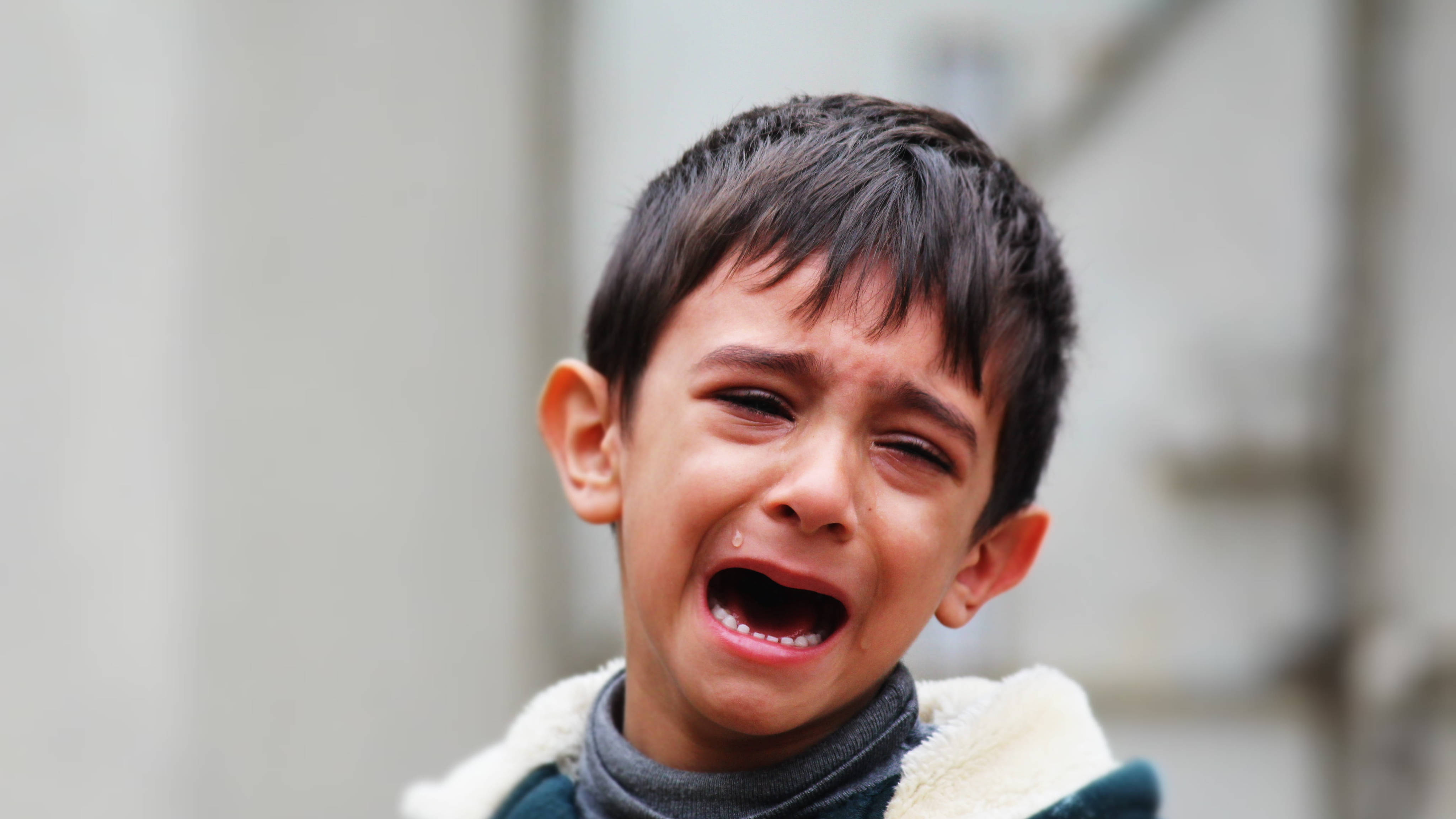 crying sad boy 桌布 壁紙 — 一個黑髮小男孩在模糊的城市背景下激烈地哭泣，淚水順著臉頰流下。