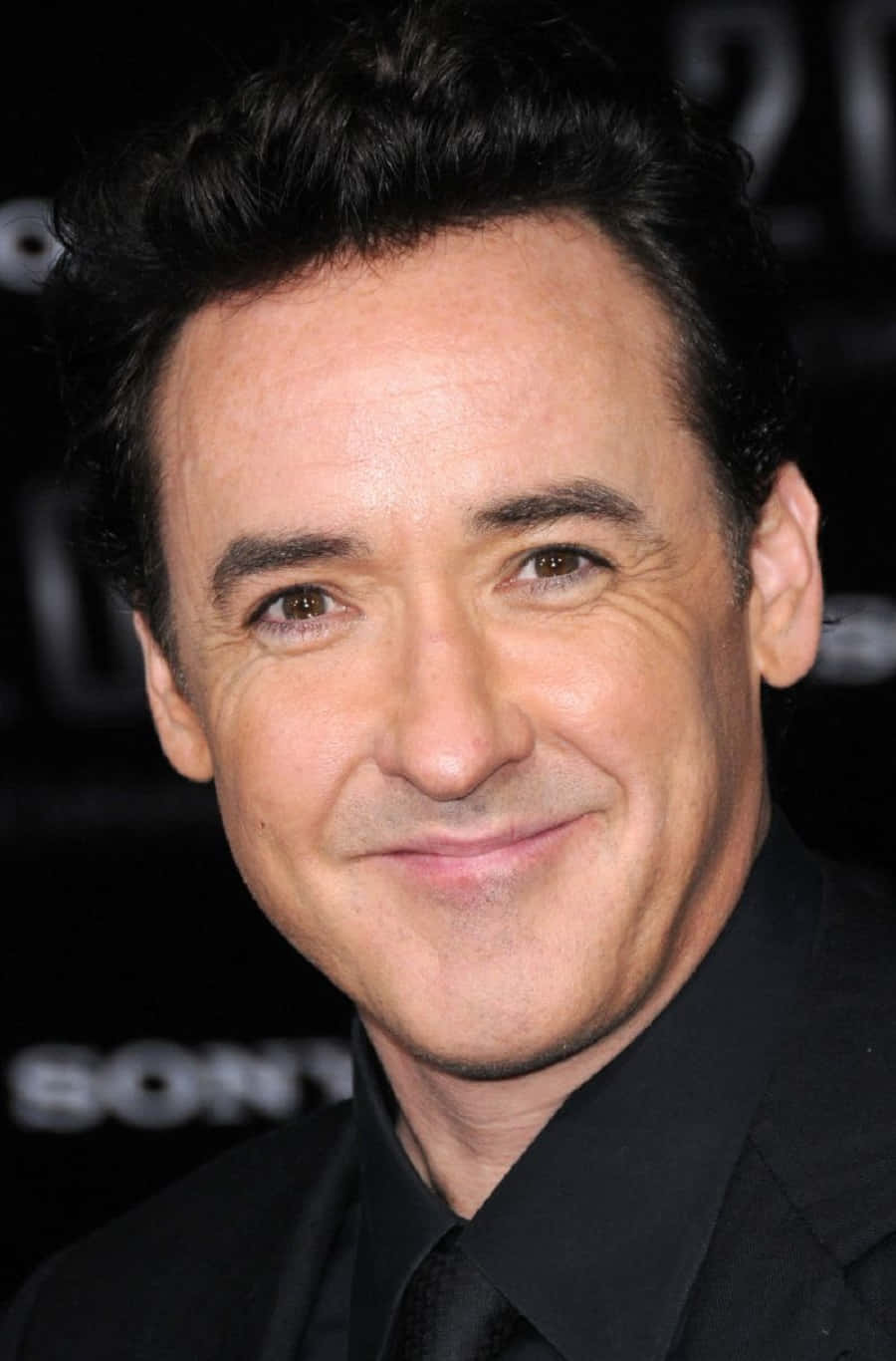 John Cusack 桌布 壁紙 — 一個黑髮微笑男子穿著黑色西裝在深色背景前的特寫肖像。