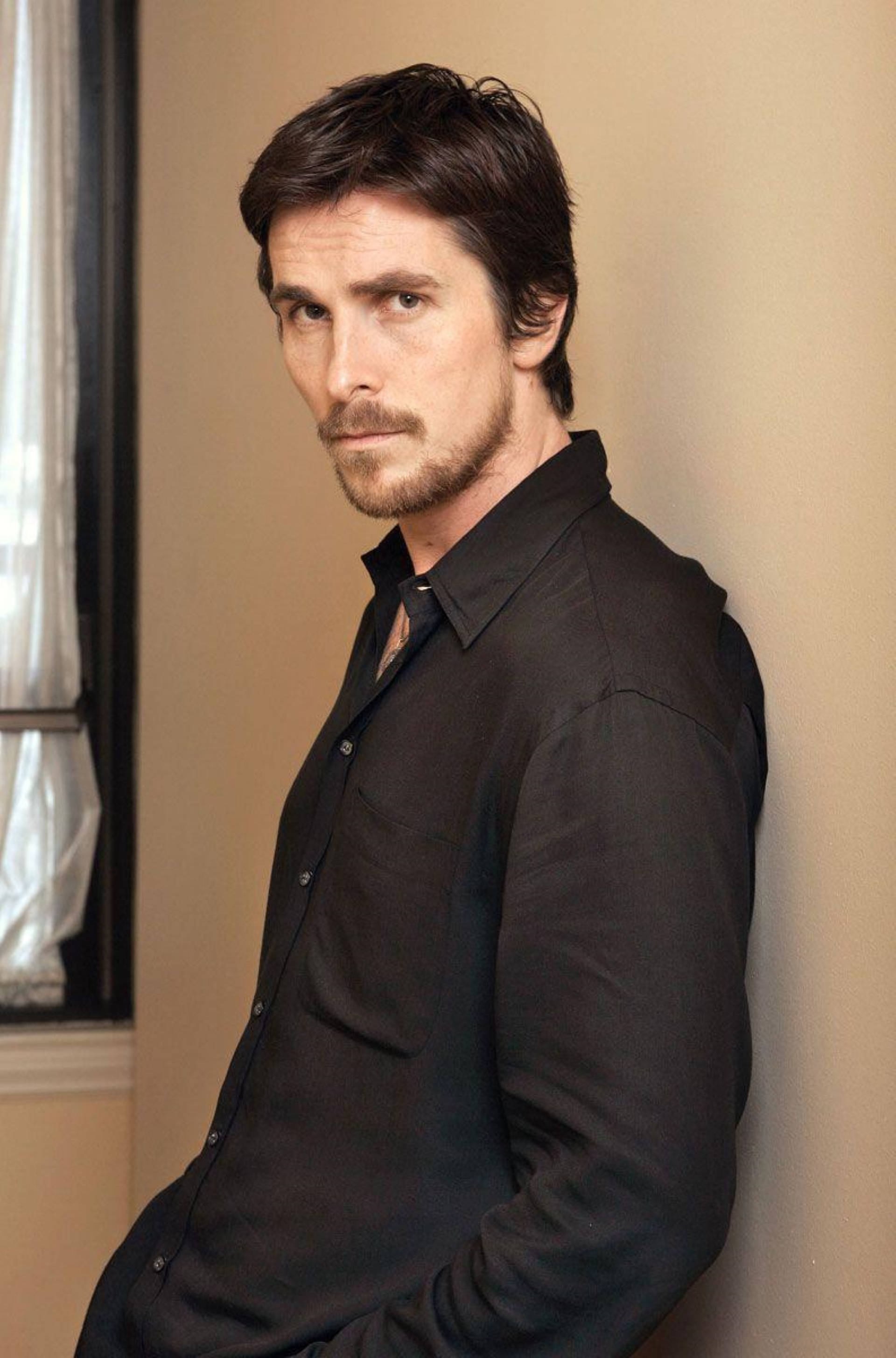 Christian Bale 桌布 壁紙 — 一個黑髮鬍鬚男靠著米色牆壁，身穿黑色鈕扣襯衫。