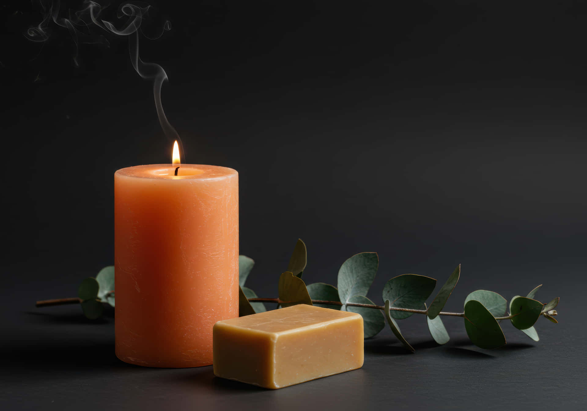 Burning candle and soap with eucalyptus on dark background 壁紙 — 一個點燃的橙色蠟燭冒著上升的煙霧，旁邊放著一塊肥皂和尤加利樹枝在黑暗的表面上。