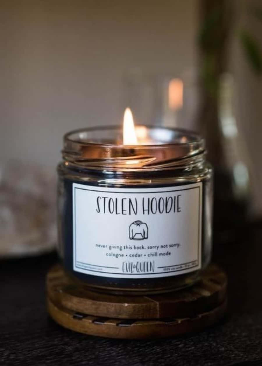 Candle Light 桌布 壁紙 — 一個點燃的玻璃蠟燭，標有「STOLEN HOODIE」，放在一個木製杯墊上，處於柔和光線的舒適室內環境中。
