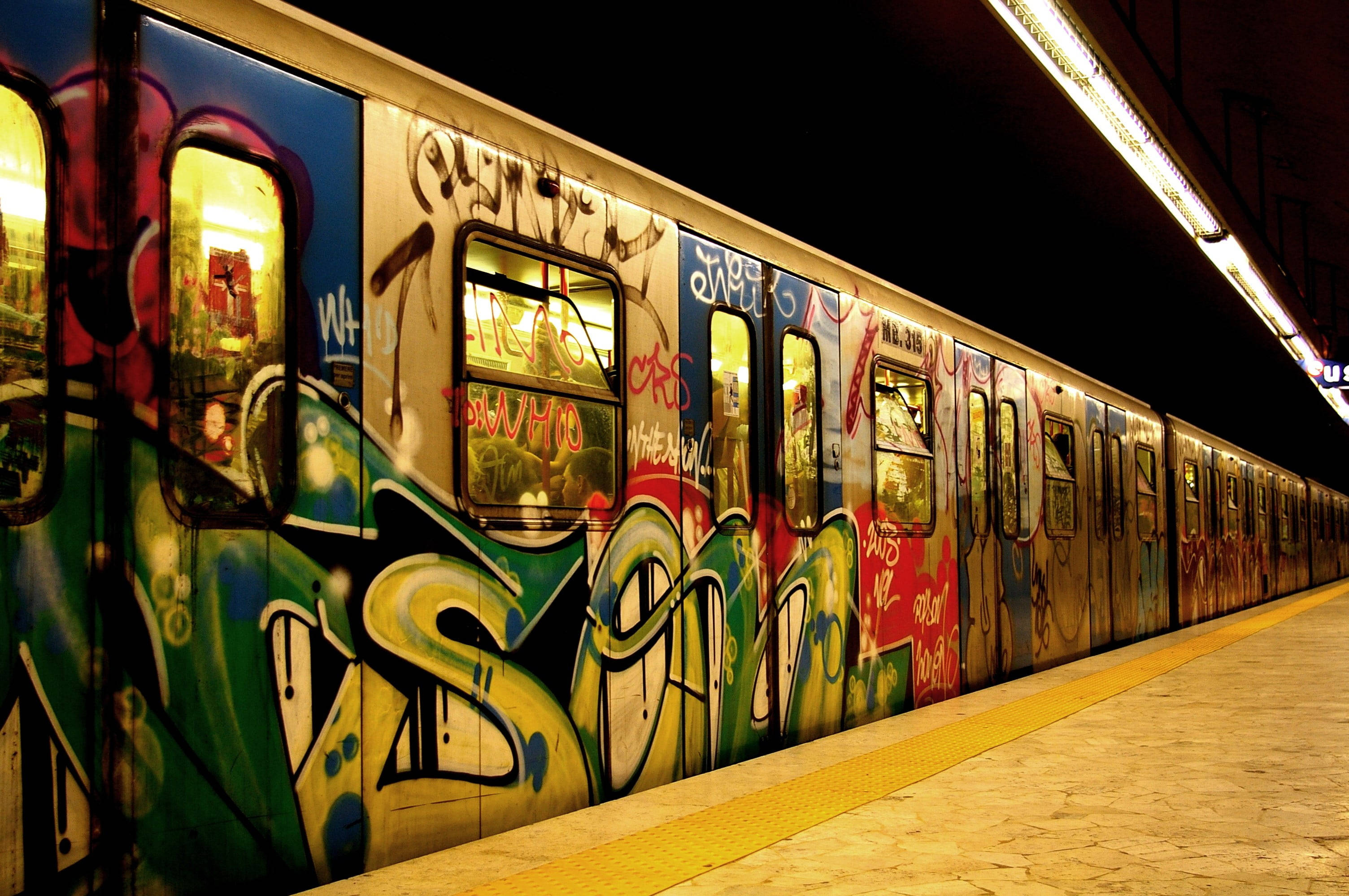 Subway 桌布 壁紙 — 一列充滿活力的塗鴉地鐵列車在夜間車站燈光下閃耀，其色彩繽紛的塗鴉標籤在黃色月台邊緣旁的窗戶中反射。