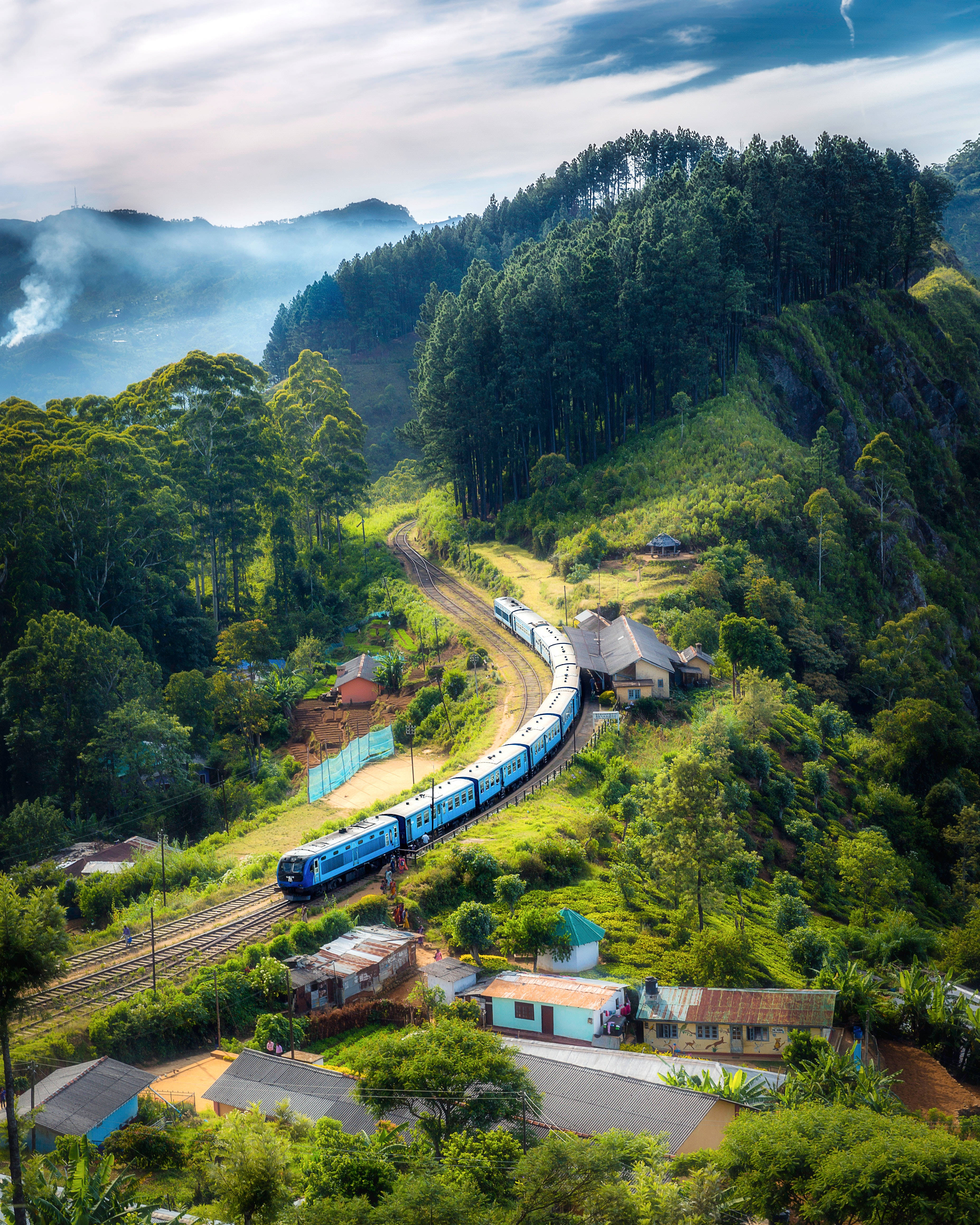 View 桌布 壁紙 — 一列充滿活力的藍色火車蜿蜒穿過翠綠的山巒，經過小村莊的房屋，天空部分多雲。