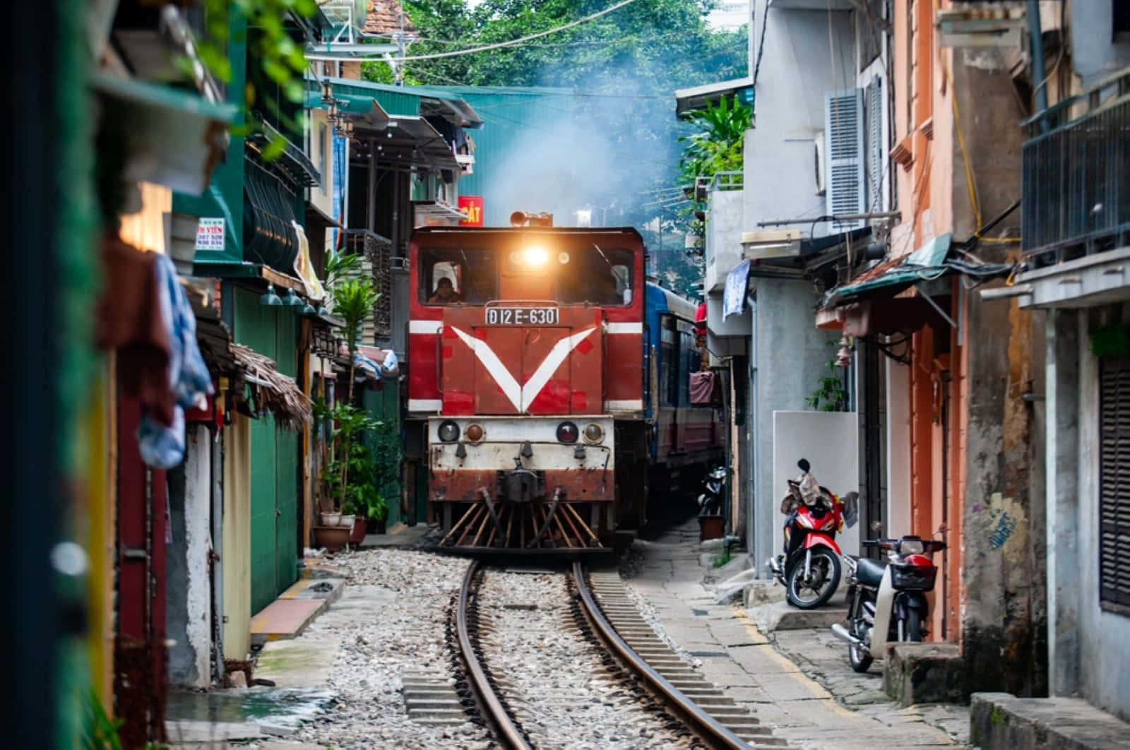 攝影 桌布 壁紙 — 一列帶有白色V標誌的紅色火車，穿過一條狹窄、飽經風霜的都市小巷，兩旁是色彩繽紛的建築物和停放的機車。