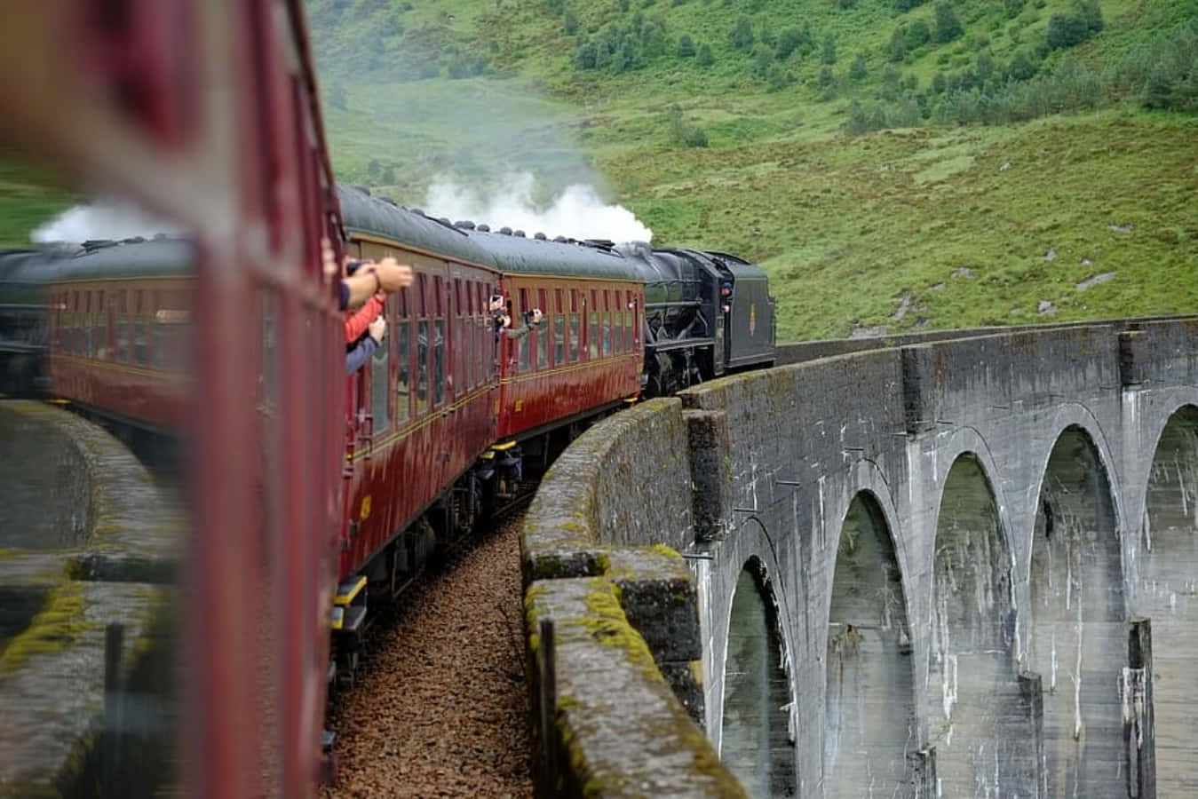 The Hogwarts Express Train 桌布 壁紙 — 一列帶有紅色車廂的復古蒸汽火車穿過長滿青苔的石拱橋，行經翠綠的山丘。