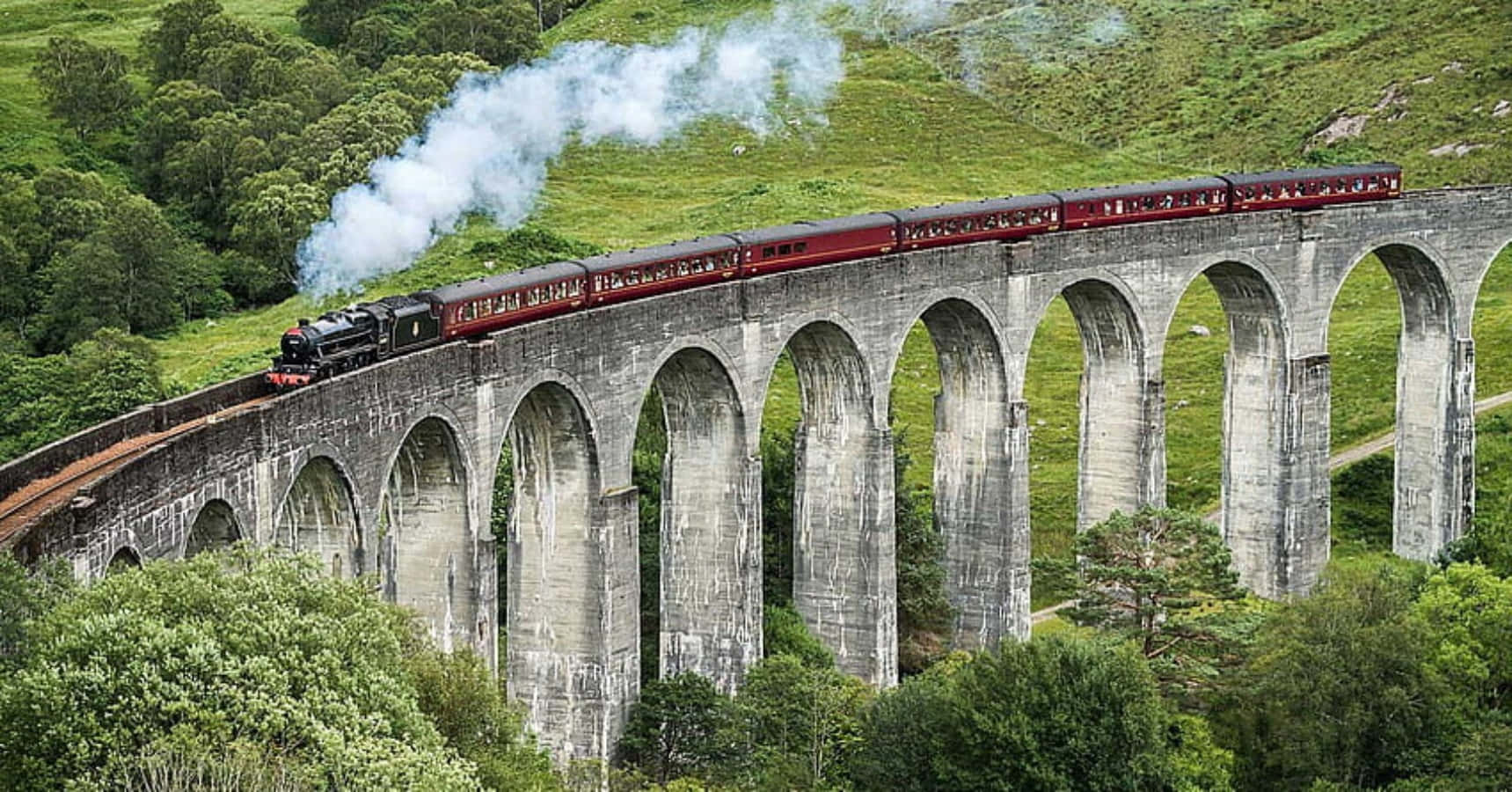 The Hogwarts Express Train 桌布 壁紙 — 一列帶有紅色車廂的蒸汽火車噴出白煙，穿過翠綠山丘中高聳的石拱橋。