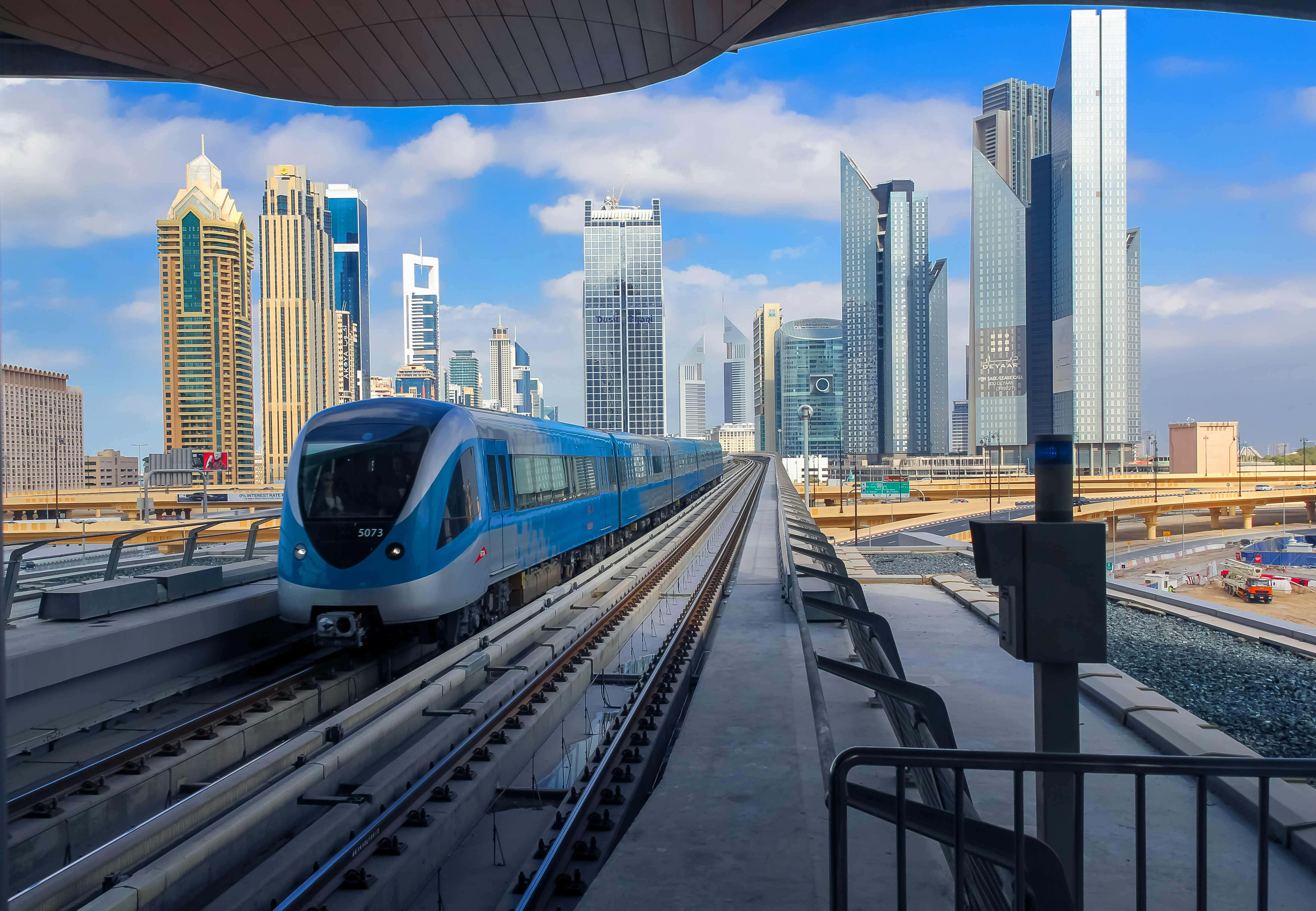 Metro 桌布 壁紙 — 一列時尚的藍色地鐵列車在現代城市天際線下沿著高架軌道滑行，玻璃摩天大樓在明亮的藍天下聳立。