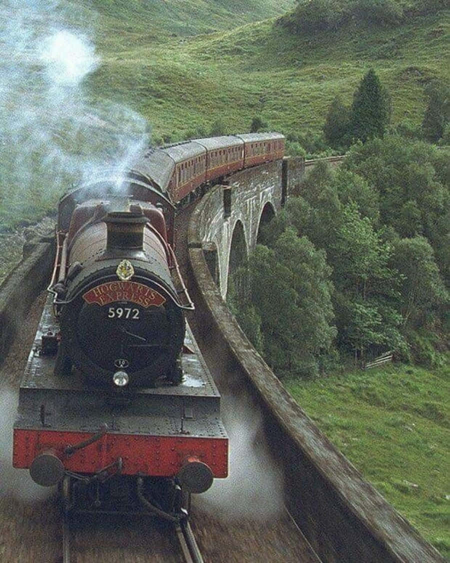 The Hogwarts Express Train 桌布 壁紙 — 一列標有「Hogwarts Express」的蒸汽火車穿過石砌高架橋，行駛在翠綠的山丘和樹木之間。