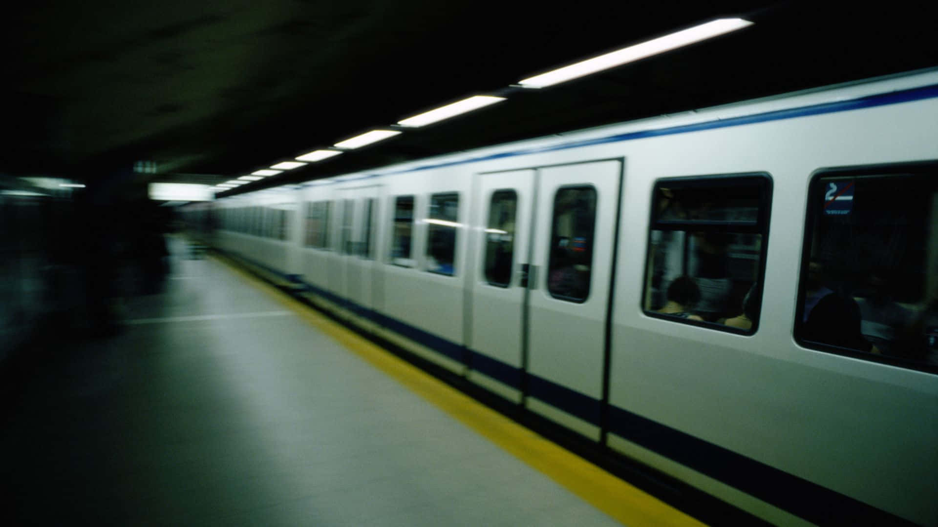 地鐵 桌布 壁紙 — 一列模糊的地鐵列車高速駛過一個燈光昏暗的月台，月台上有黃色安全線和頭頂燈光。
