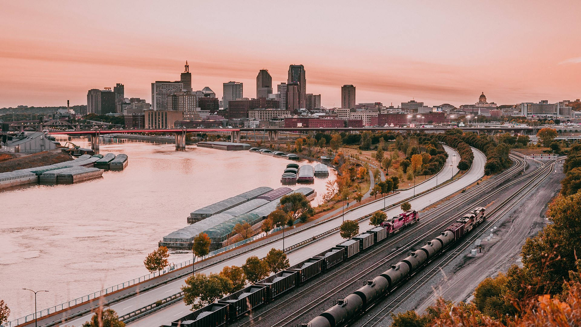 Minnesota 桌布 壁紙 — 一列火車沿著河邊的軌道行駛，城市天際線在柔和的粉紅色夕陽下閃耀。