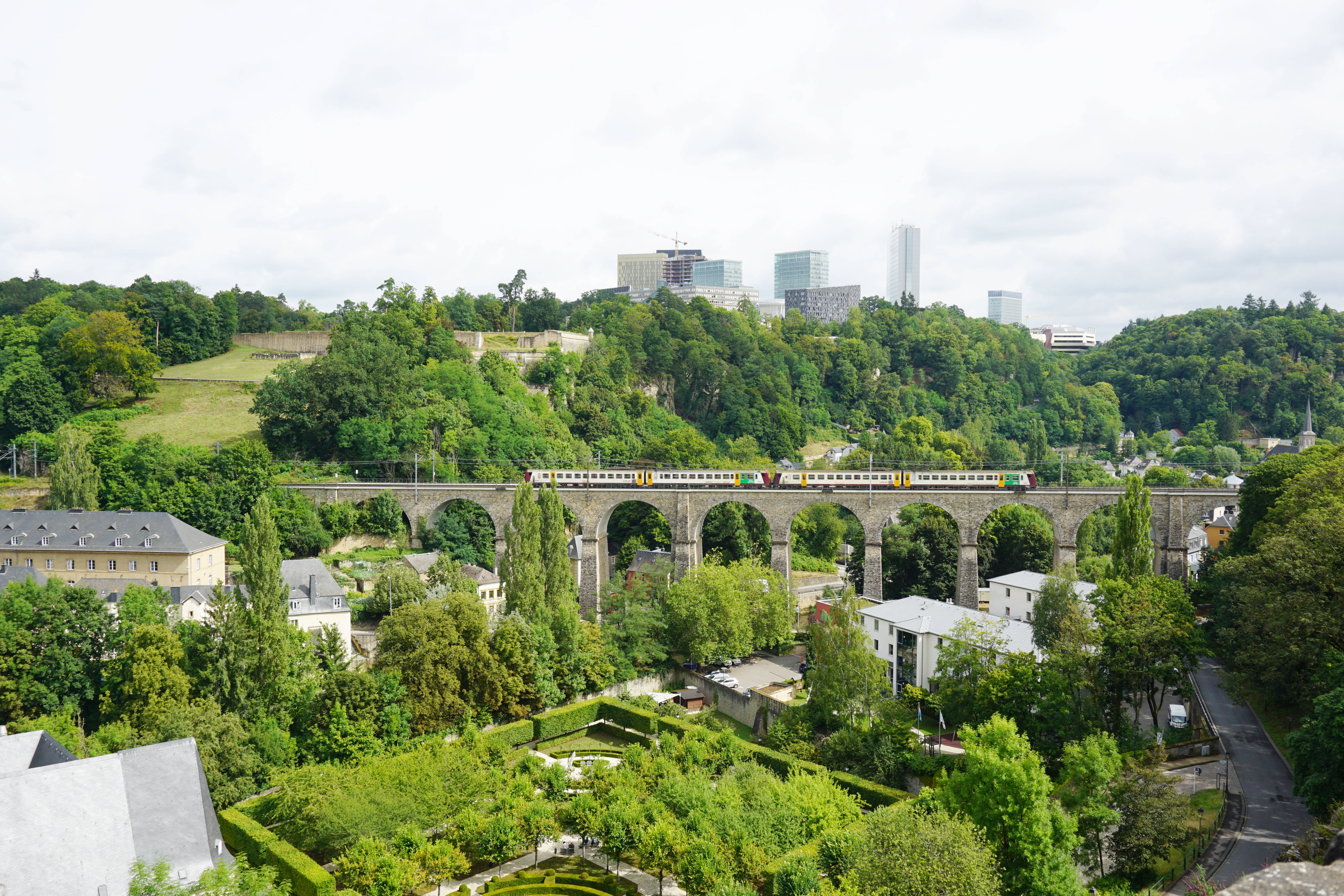 Luxembourg 桌布 壁紙 — 一列火車穿過石拱橋，下方是鬱鬱蔥蔥的花園，現代城市建築在樹木覆蓋的山丘後方升起，天空多雲。