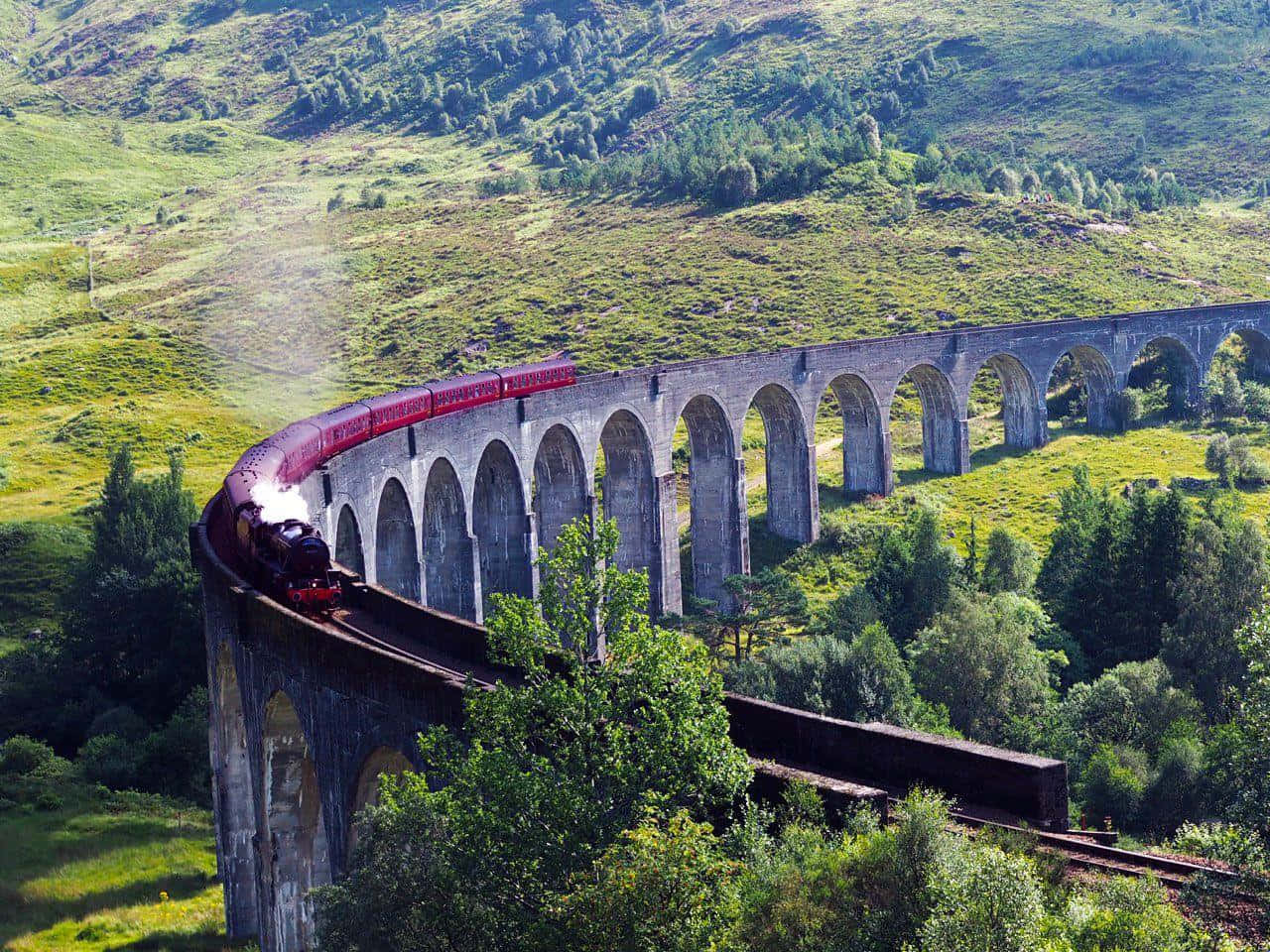 Hogwarts Express 桌布 壁紙 — 一列紅色蒸汽火車在明媚陽光下，優雅地彎曲穿過鬱蔥的綠色山丘，行駛在一座高聳的石拱橋上。