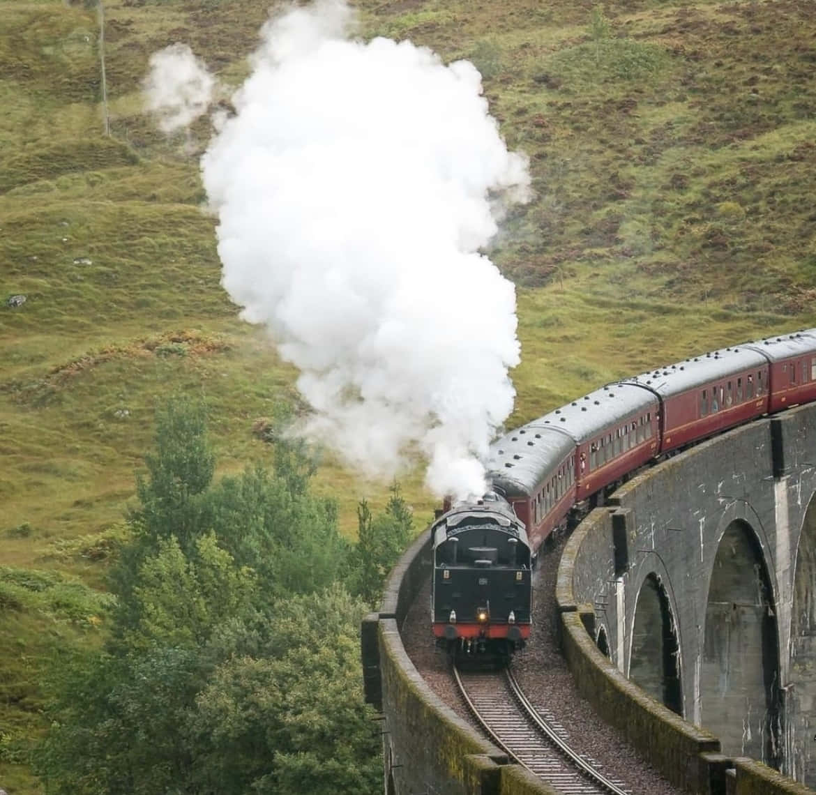 Hogwarts Express 桌布 壁紙 — 一列紅色車廂的蒸汽火車穿過石砌高架橋，穿越翠綠的山丘，噴出濃厚的白煙。