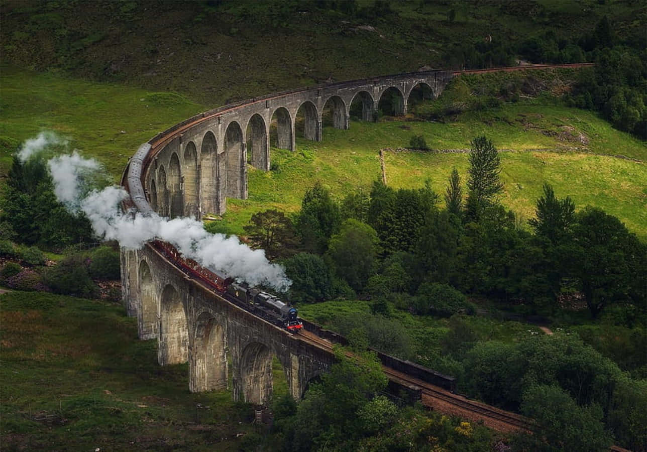 The Hogwarts Express Train 桌布 壁紙 — 一列蒸汽火車噴出白煙，沿著宏偉的石拱橋蜿蜒穿過鬱鬱蔥蔥的綠色山丘和茂密的森林。
