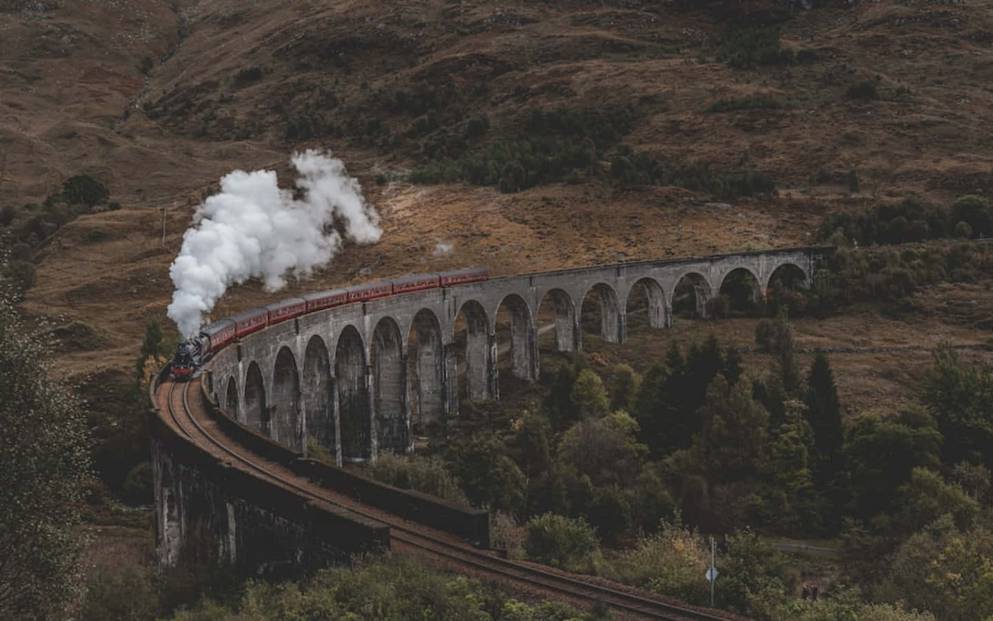 The Hogwarts Express Train 桌布 壁紙 — 一列蒸汽火車噴出白煙，沿著蜿蜒的石拱橋穿過崎嶇、樹木點綴的山丘行駛。