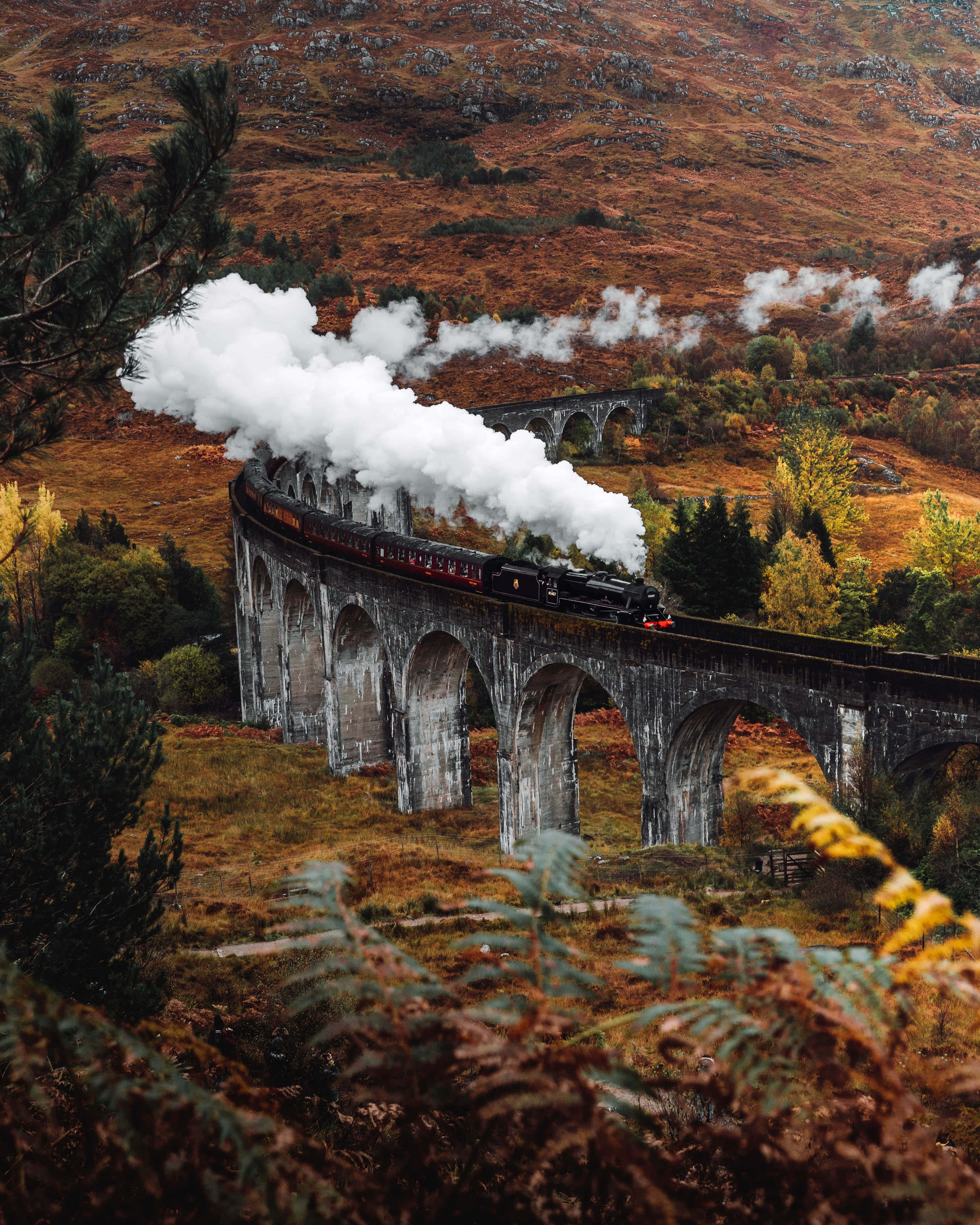 Hogwarts Express 桌布 壁紙 — 一列蒸汽火車噴出白煙，穿越崎嶇秋日山谷中的高聳石橋。