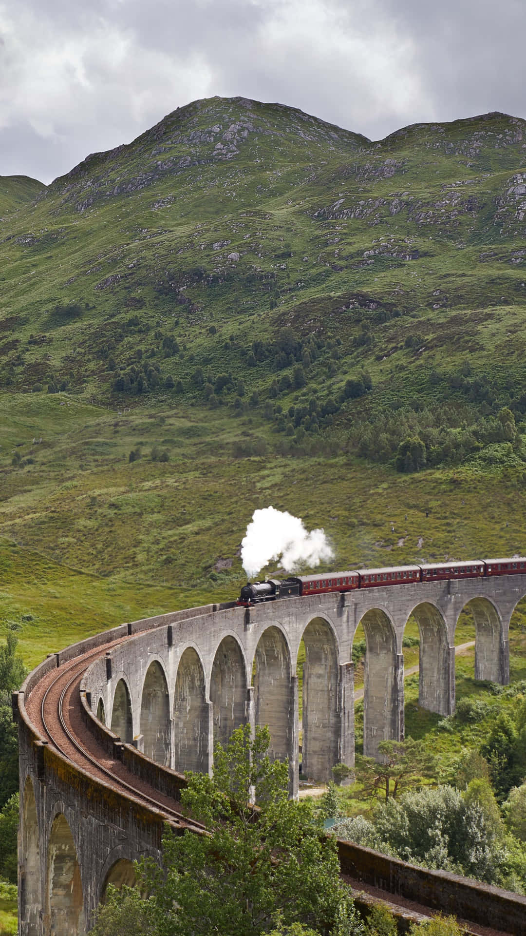 Hogwarts Express 桌布 壁紙 — 一列蒸汽火車噴出白煙，穿過一條宏偉的石砌高架橋，蜿蜒於蒼翠的山丘之間，天空多雲。
