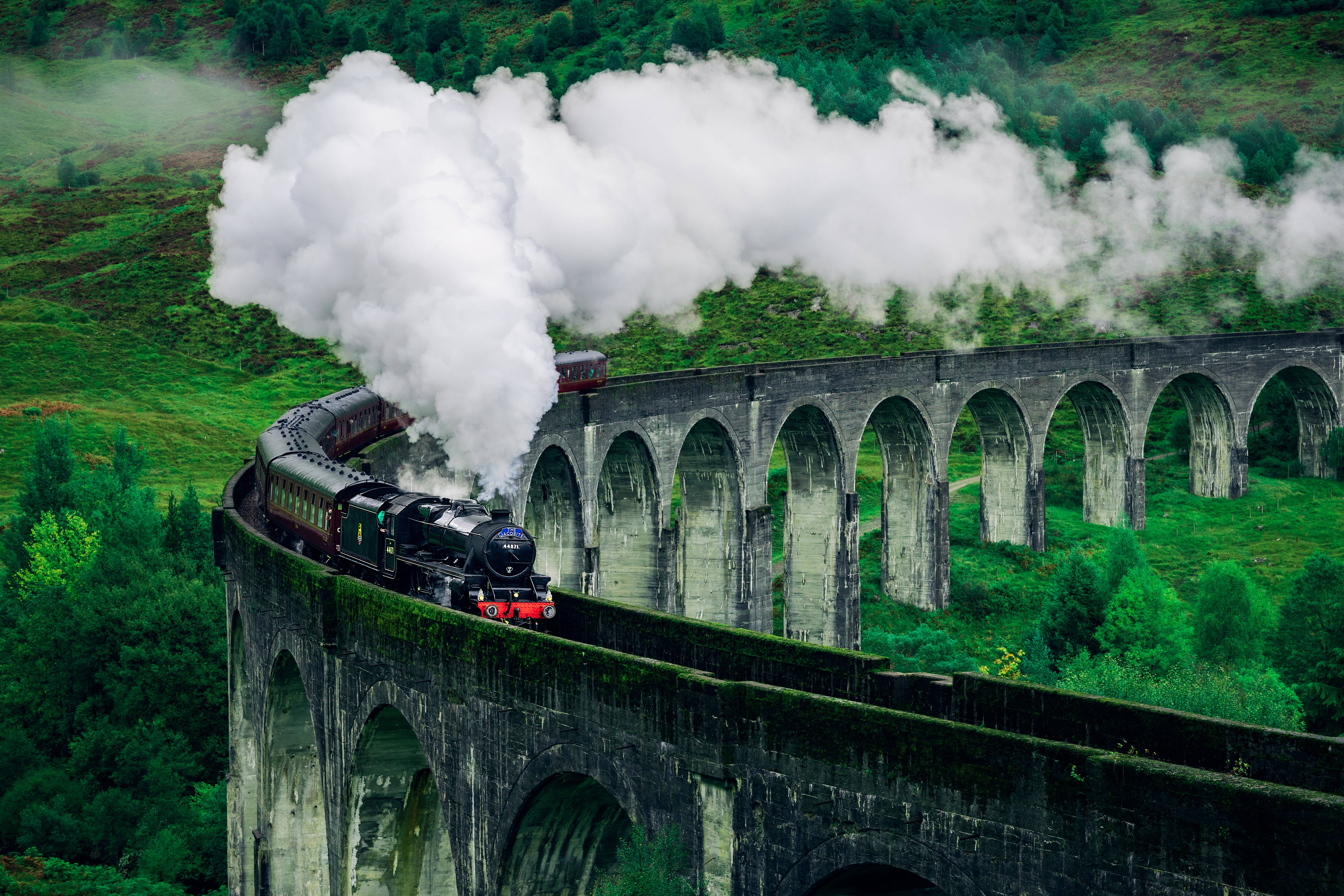 Train 桌布 壁紙 — 一列蒸汽火車沿著高聳的石拱橋穿過鬱鬱蔥蔥的綠色山丘，噴出濃厚的白煙。