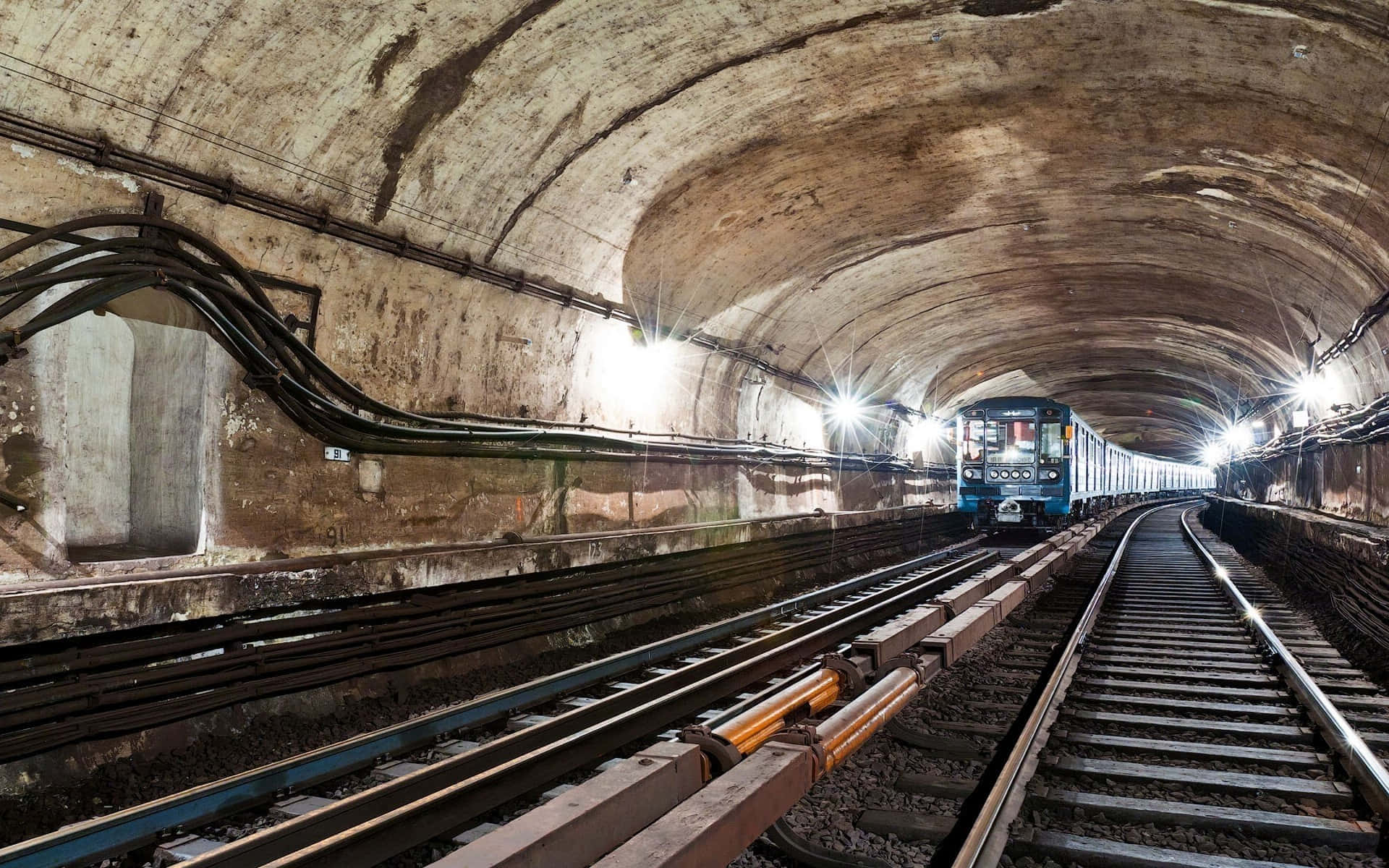 Metro 桌布 壁紙 — 一列藍色地鐵列車正穿過一條燈光昏暗、拱形混凝土隧道，隧道內有外露管道和發光的頂燈。