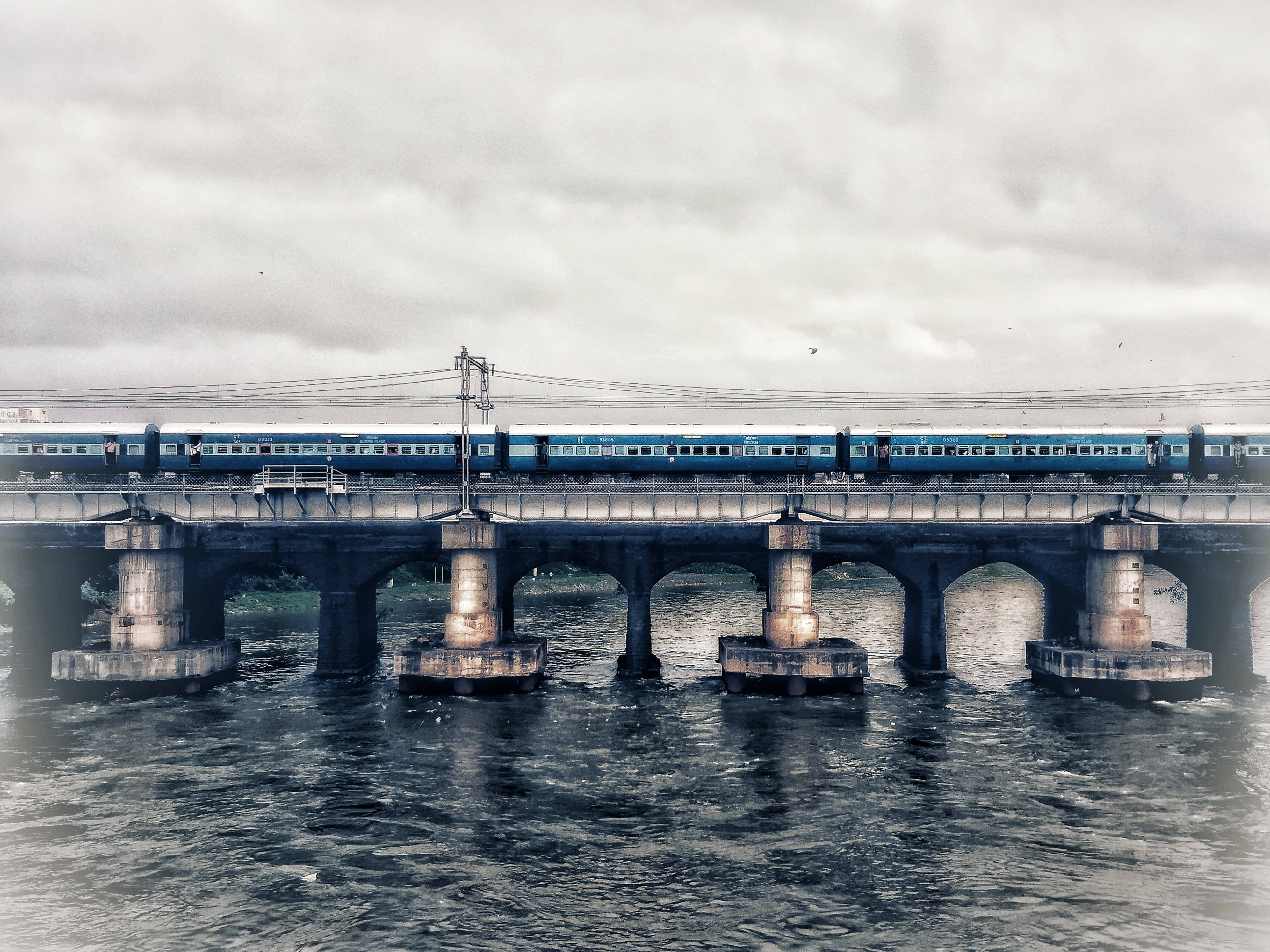 Mumbai 桌布 壁紙 — 一列藍色客運列車在陰天下穿越一座風化的石橋，橋下波濤洶湧。