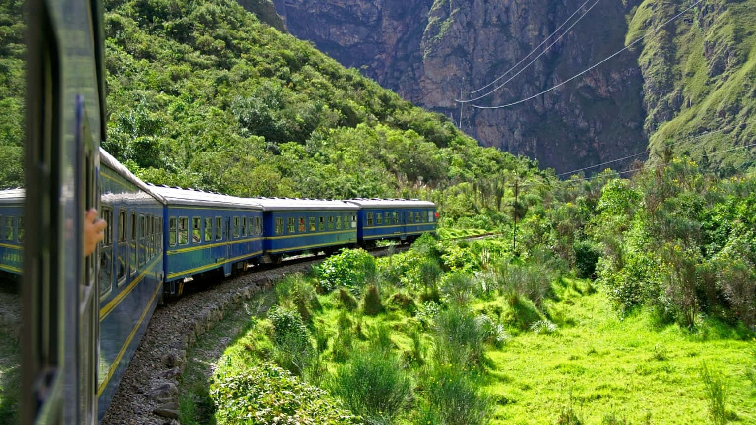 1536x864 桌布 壁紙 — 一列藍色火車蜿蜒穿過鬱鬱蔥蔥的綠色峽谷，兩旁是陡峭的森林懸崖，在明亮的日光下。