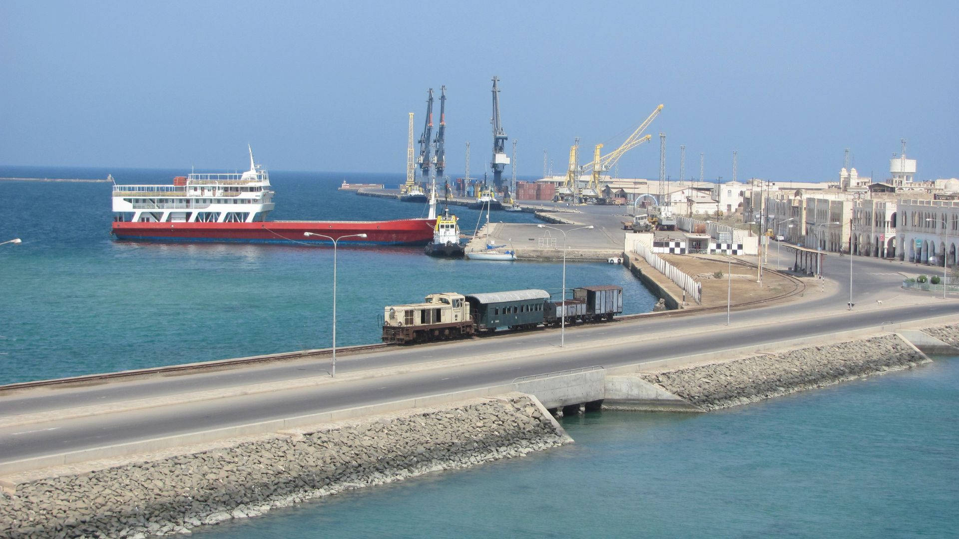 Eritrea 桌布 壁紙 — 一列貨運火車正駛過海邊橋樑，一艘紅色貨船在晴朗藍天下停泊於繁忙港口。