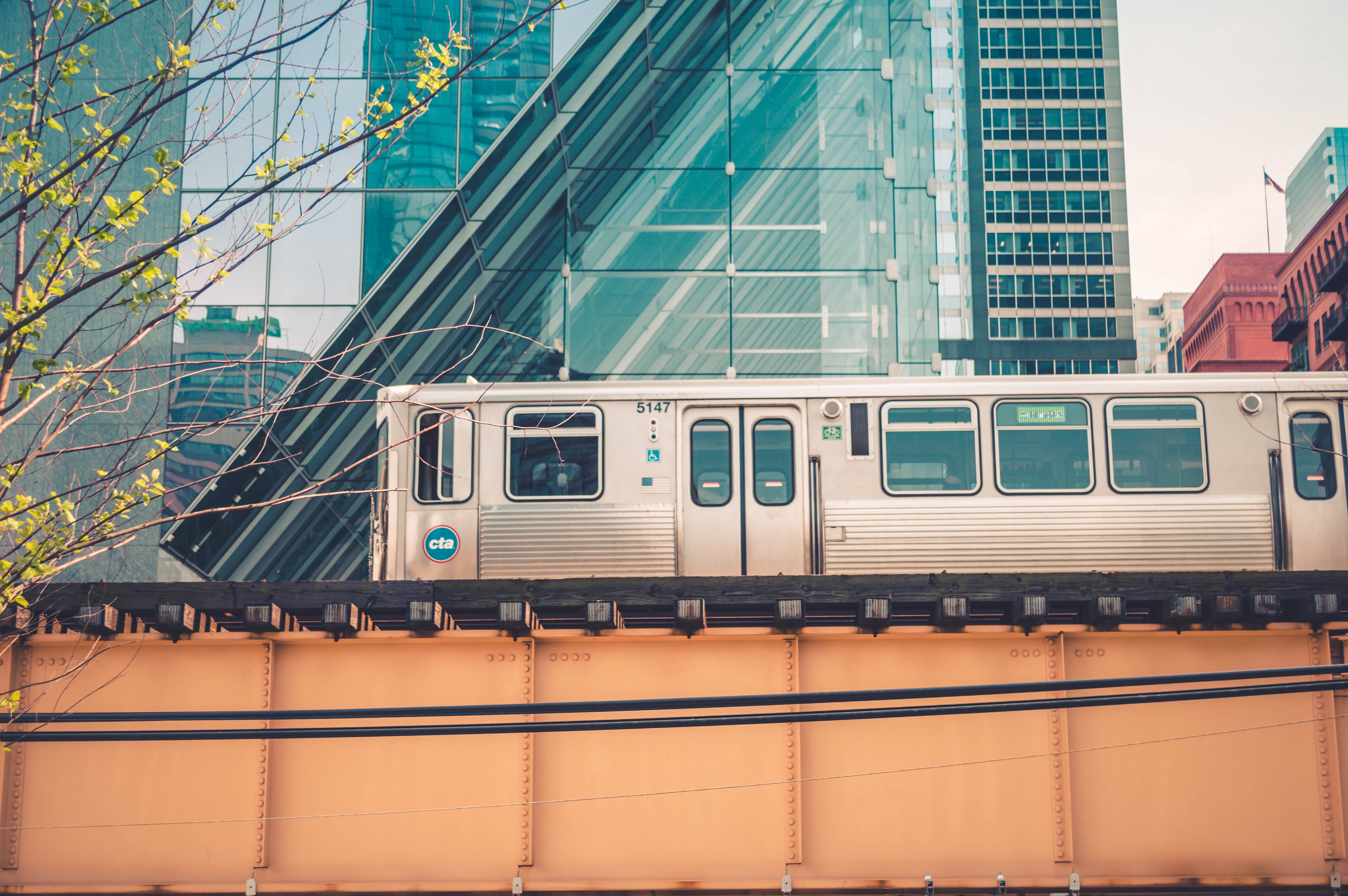 火車 桌布 壁紙 — 一列銀色火車在橙色橋上行駛，橋下是玻璃摩天大樓，前景有嫩綠的樹枝。