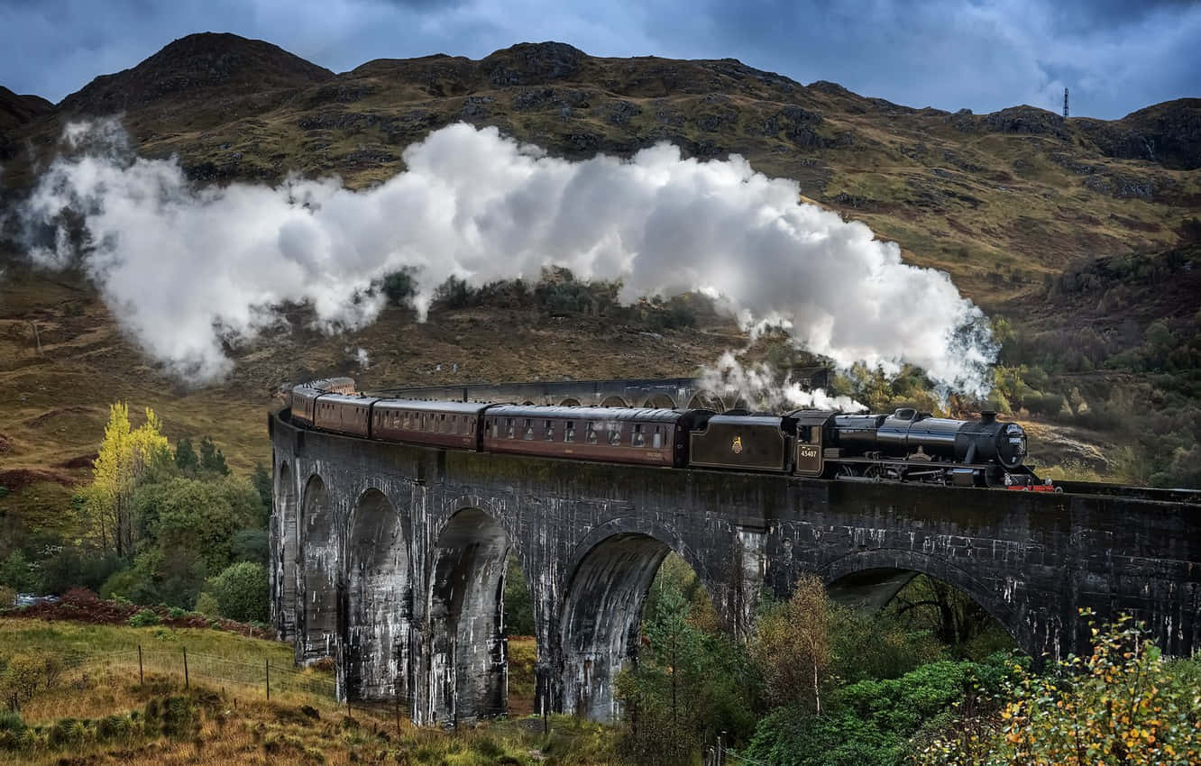 Hogwarts Express 桌布 壁紙 — 一列雄偉的蒸汽火車噴出濃厚的白煙，穿越崎嶇的秋日山丘，行駛在一座高大的石拱橋上。
