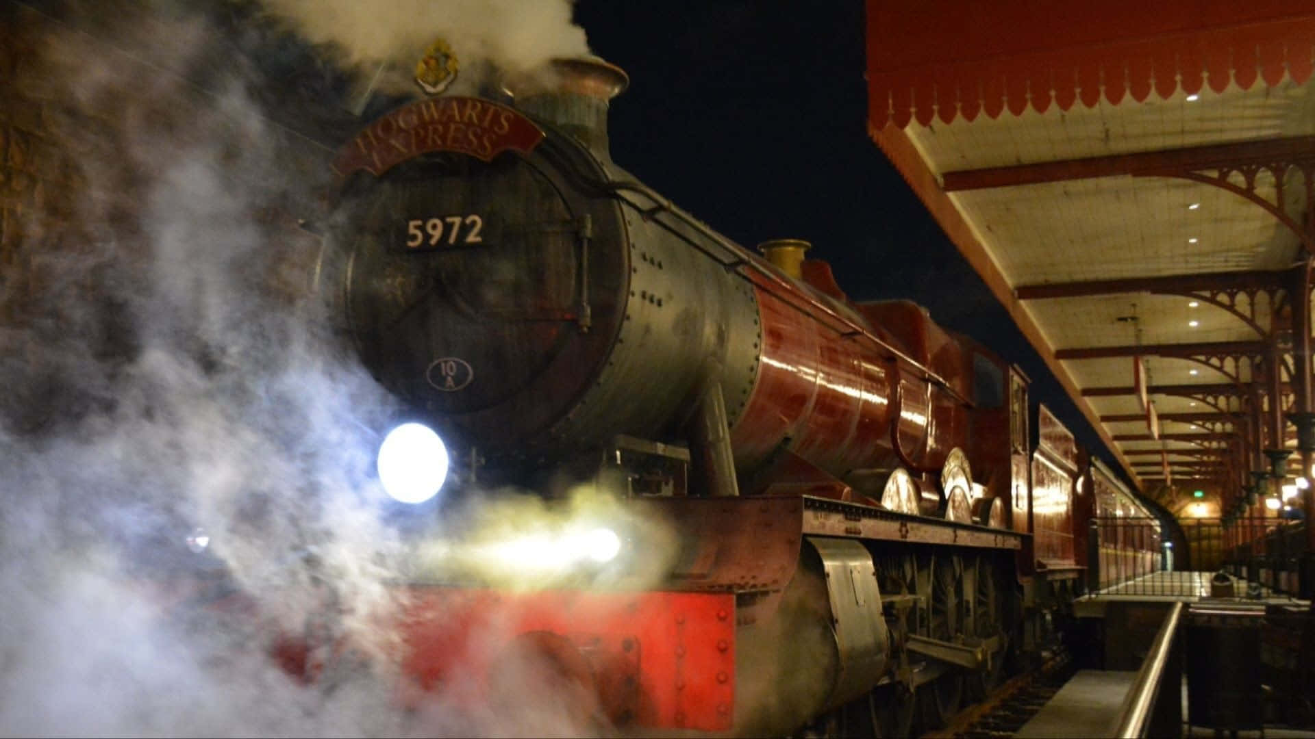 Hogwarts Express 桌布 壁紙 — 一列雄偉的霍格華茲特快蒸汽火車在夜間冒著煙駛入一個大型車站月台。