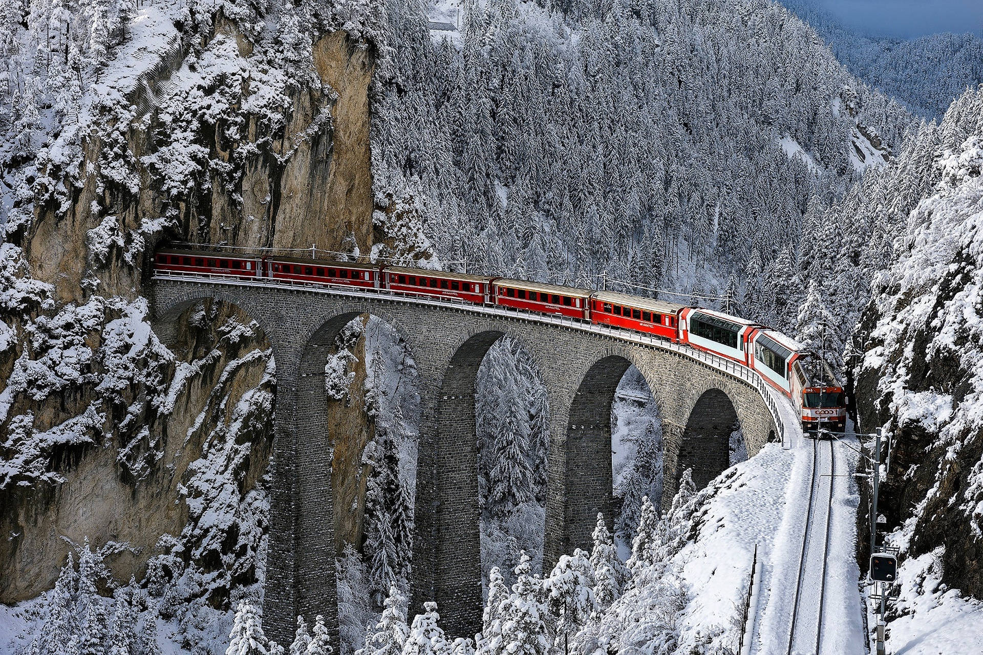 Alps 桌布 壁紙 — 一列鮮紅色的火車優雅地彎曲穿過白雪覆蓋的阿爾卑斯森林，越過高聳的石橋。