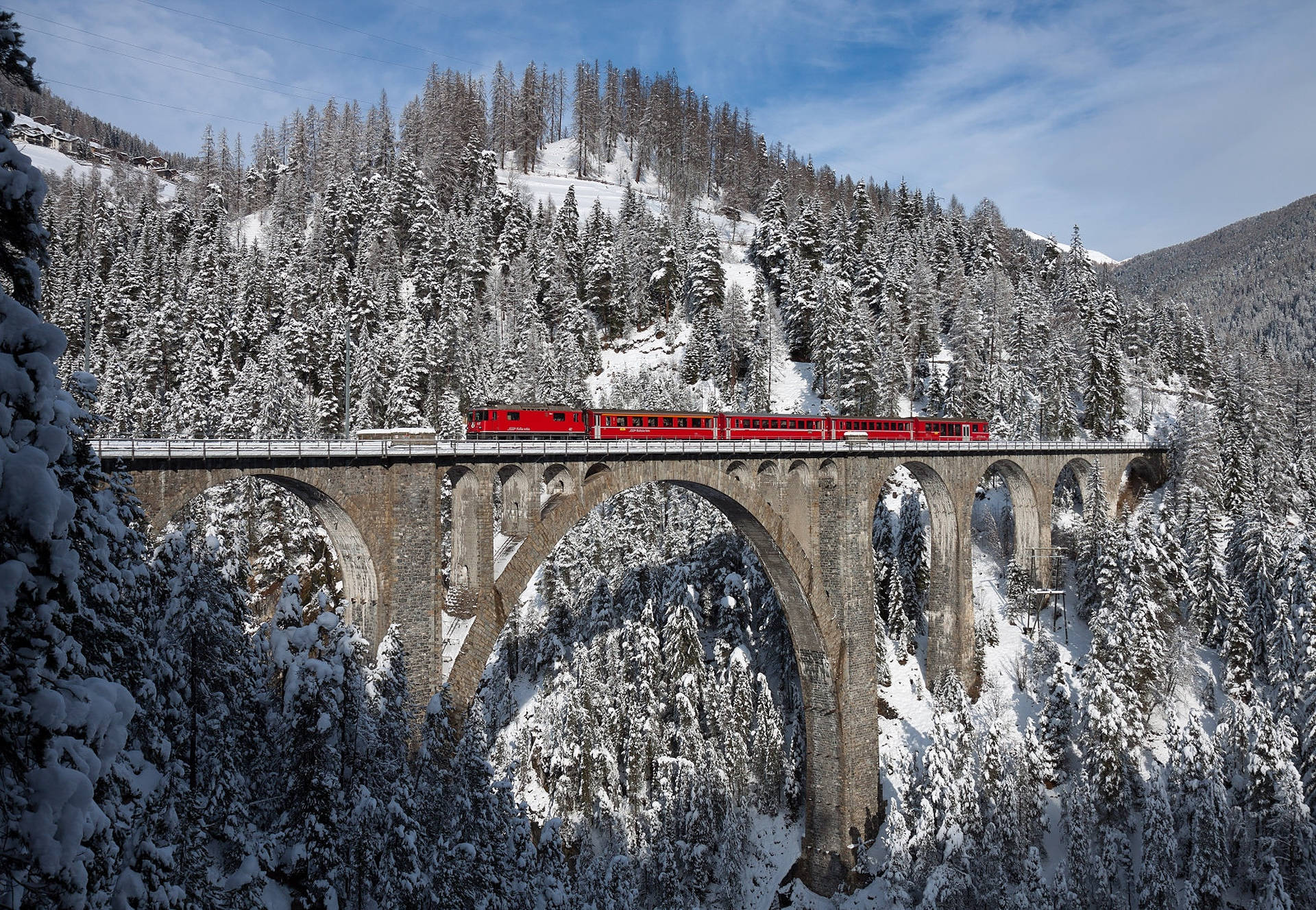 Alps 桌布 壁紙 — 一列鮮紅色的火車穿過一座宏偉的石拱橋，行經白雪覆蓋的森林山谷，在清澈的藍天下。