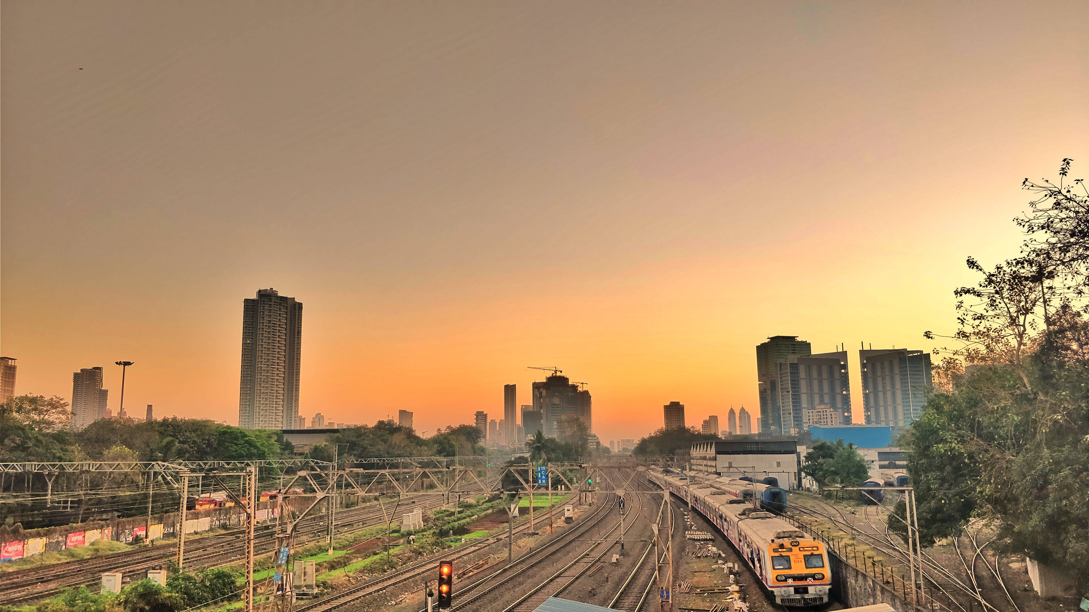 Mumbai 桌布 壁紙 — 一列黃色火車在日落時分滑行穿過廣闊的城市鐵路調車場，高聳的摩天大樓在溫暖的橙色天空下呈現剪影。
