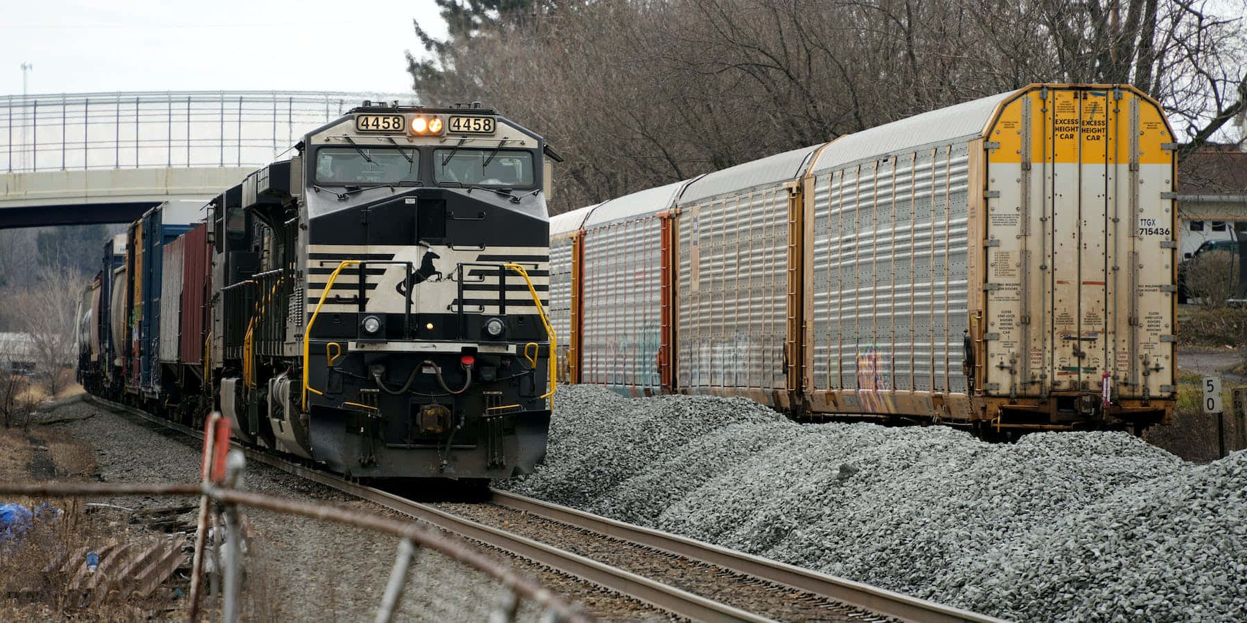 火車 桌布 壁紙 — 一列黑色和黃色的貨運火車，編號4458，帶領一列長長的貨運列車穿過一條鋪有礫石的鐵路，經過一座鋼橋，背景是光禿禿的樹木。
