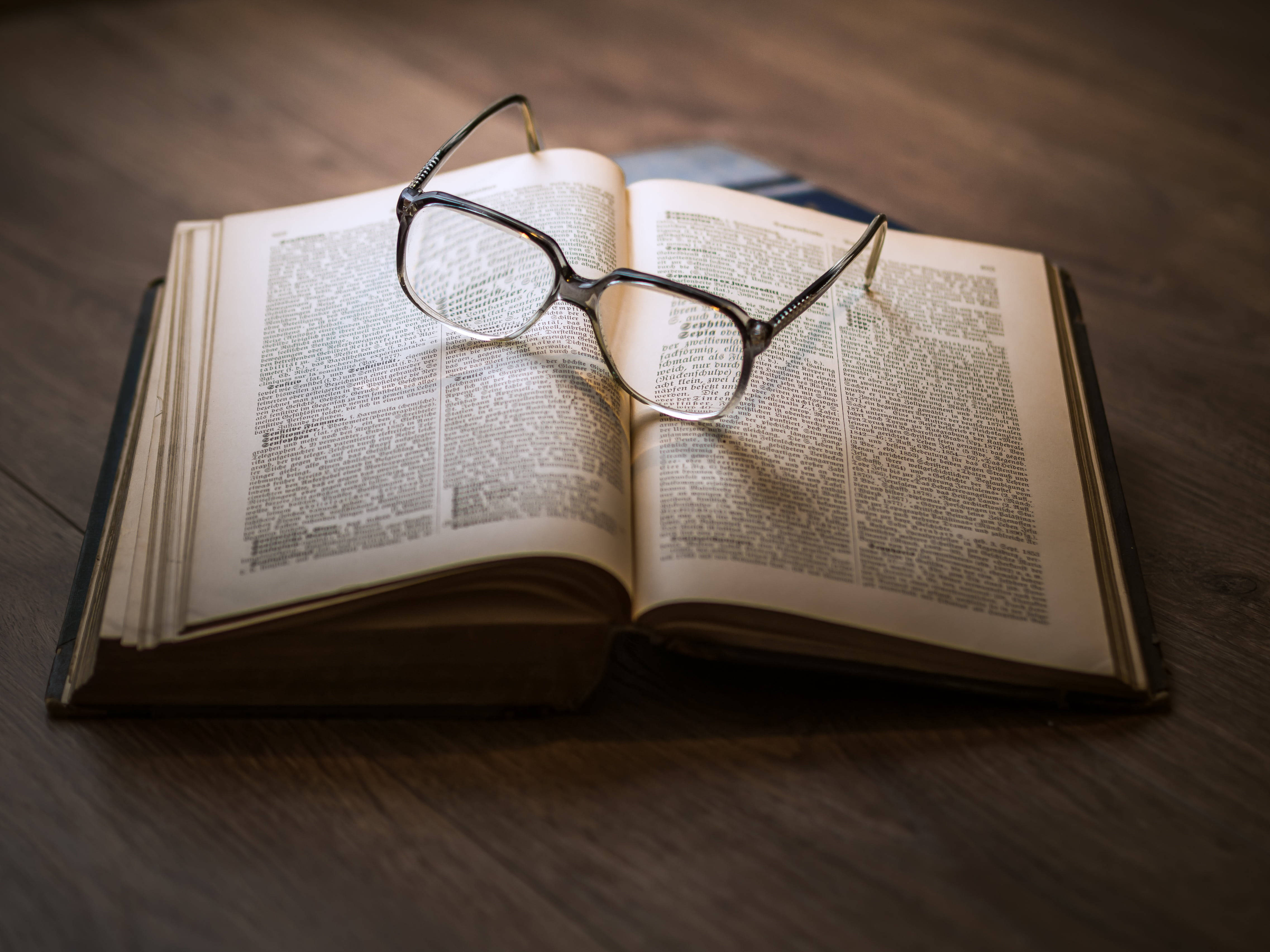 Reading 桌布 壁紙 — 一副眼鏡擱在打開的字典上，字典放在木質表面上，光線溫暖柔和。