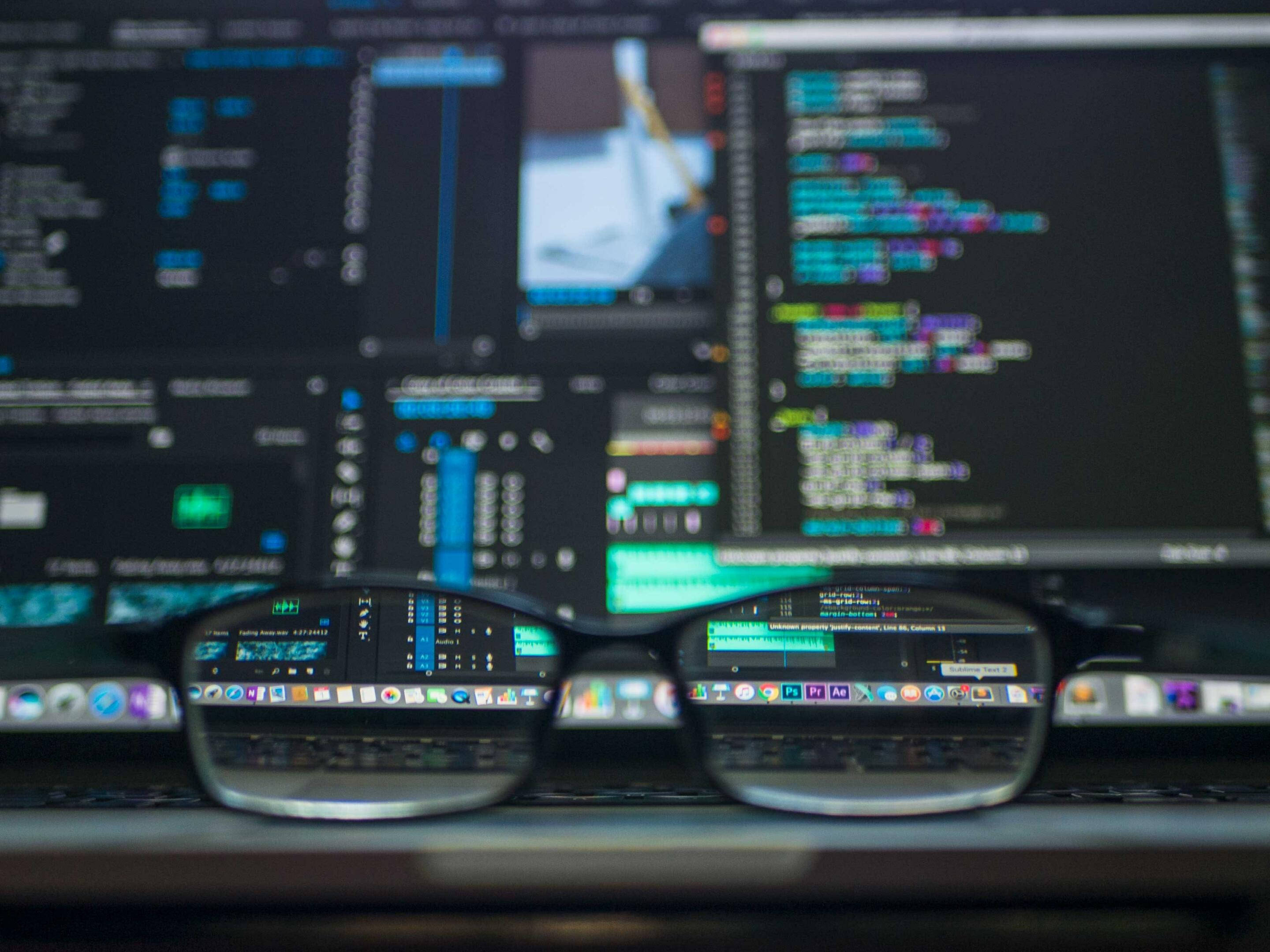 Code 桌布 壁紙 — 一副眼鏡放在鍵盤上，背景是模糊的螢幕，顯示色彩繽紛的程式碼和設計軟體。