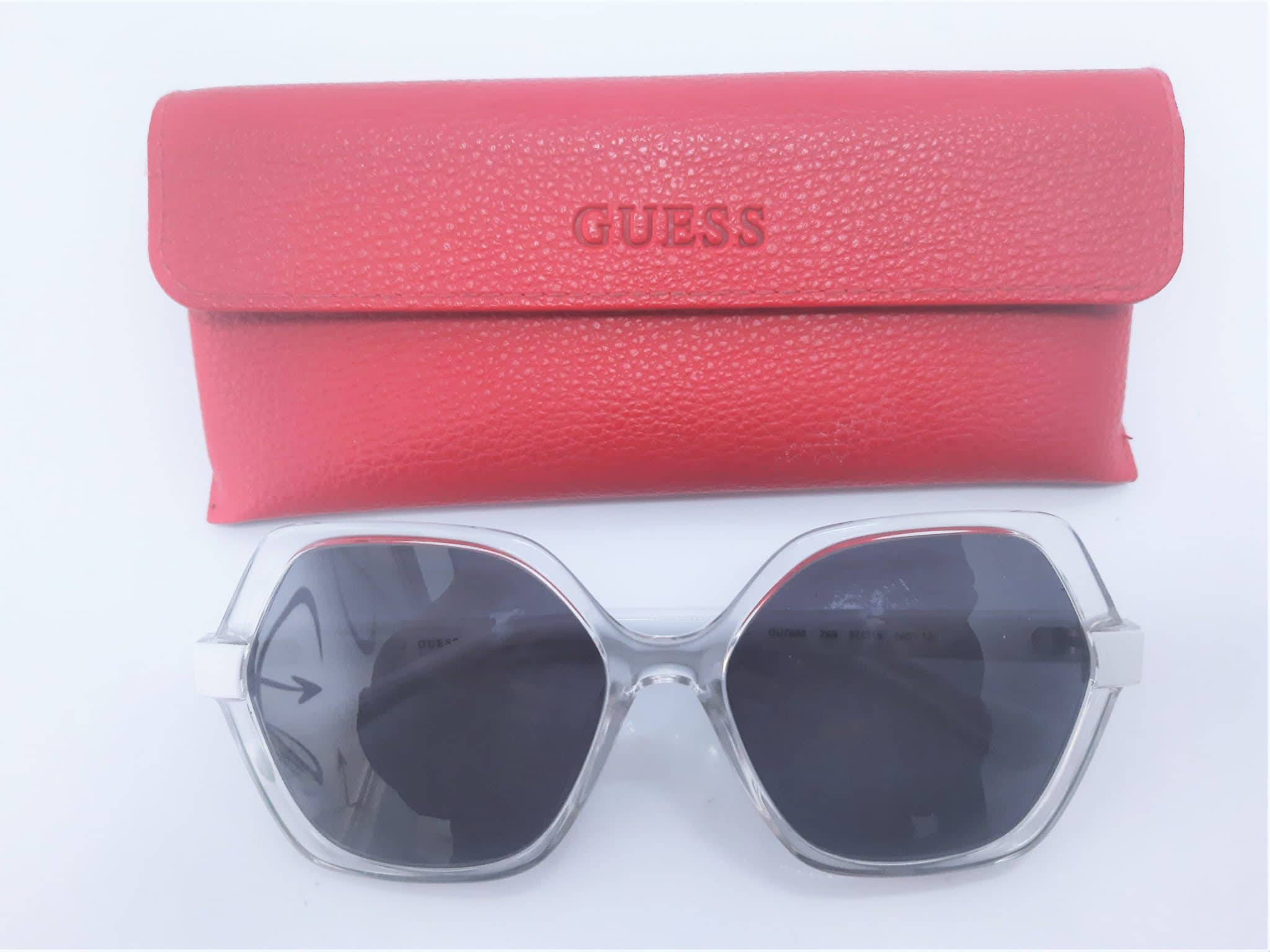 Sunglasses 桌布 壁紙 — 一副透明鏡框的Guess太陽眼鏡放在一個有紋理的珊瑚粉紅色皮革盒下方，背景是潔淨的白色。