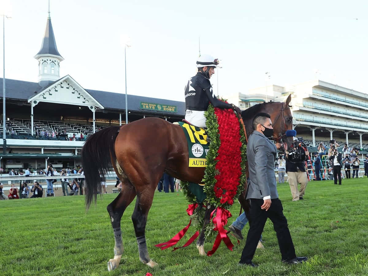 Kentucky Derby 桌布 壁紙 — 一匹勝利的栗色賽馬，飾有紅色花環，由騎師和馬伕帶領穿過賽馬場的草地跑道。