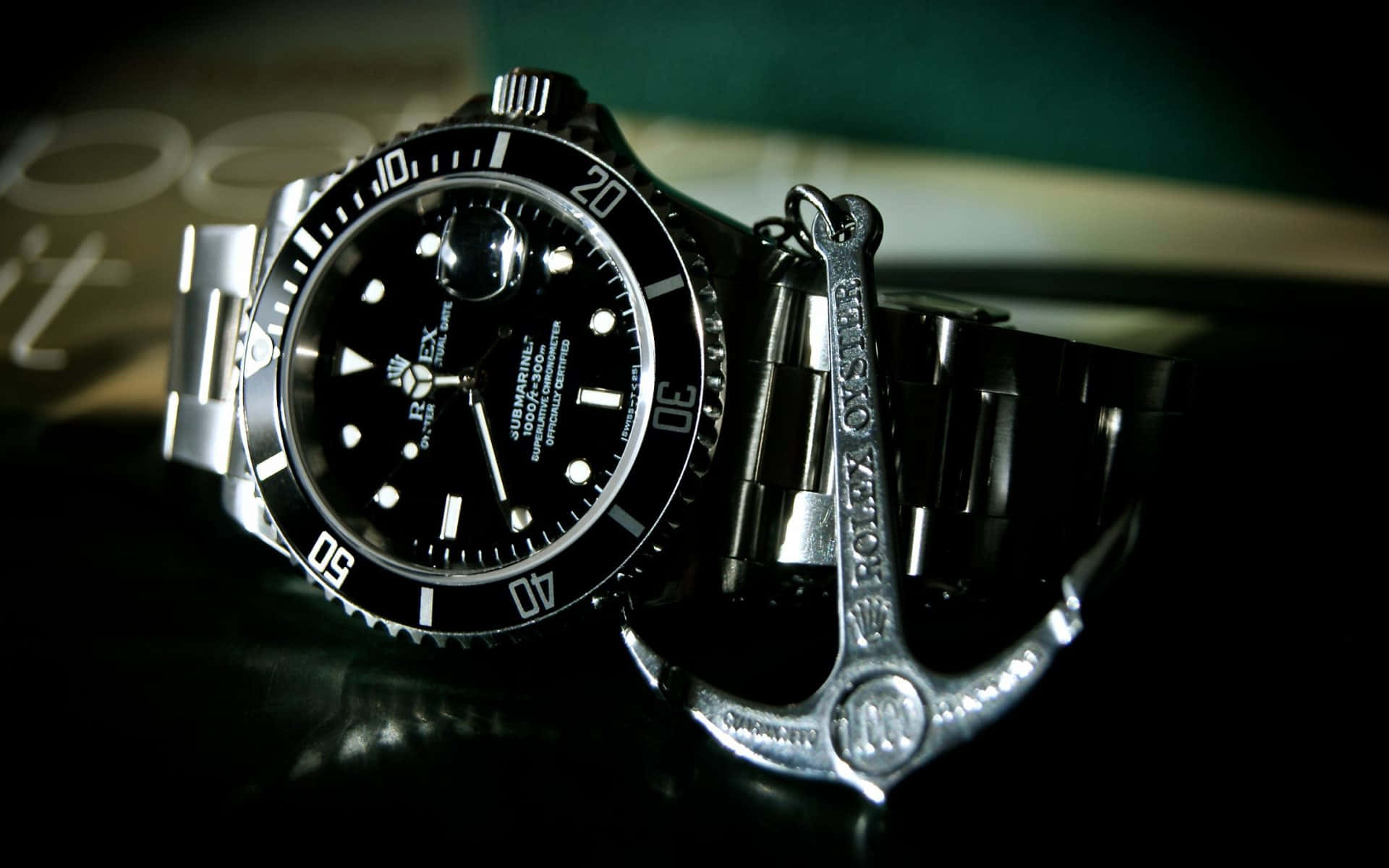 Rolex 桌布 壁紙 — 一只時尚的黑色勞力士Submariner手錶在戲劇性燈光下，於深色反光表面旁與銀色錨形鑰匙扣一同閃耀。
