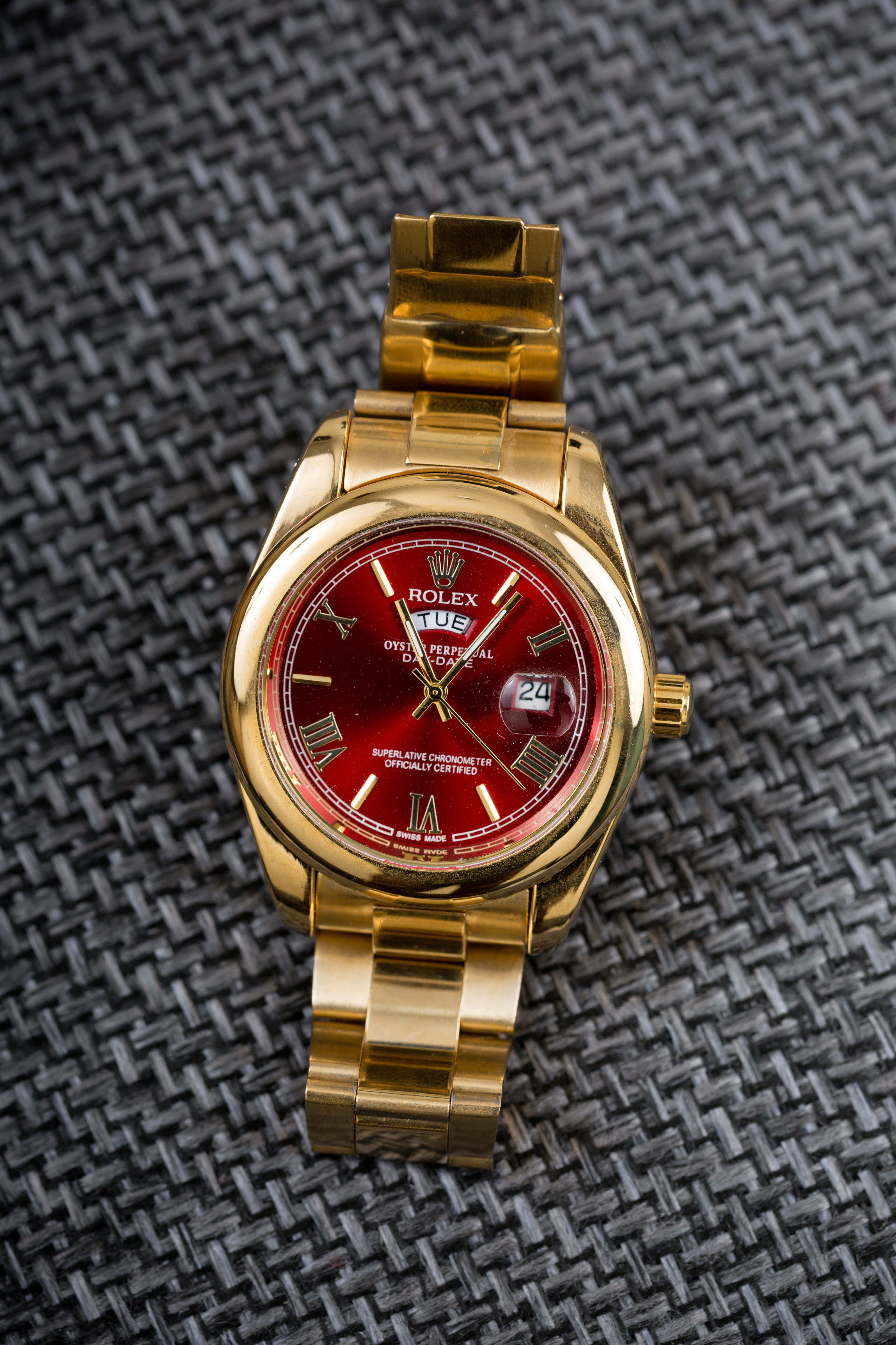 Rolex 桌布 壁紙 — 一只金色勞力士Day-Date手錶配紅色錶盤，放置在深色編織布料表面上。