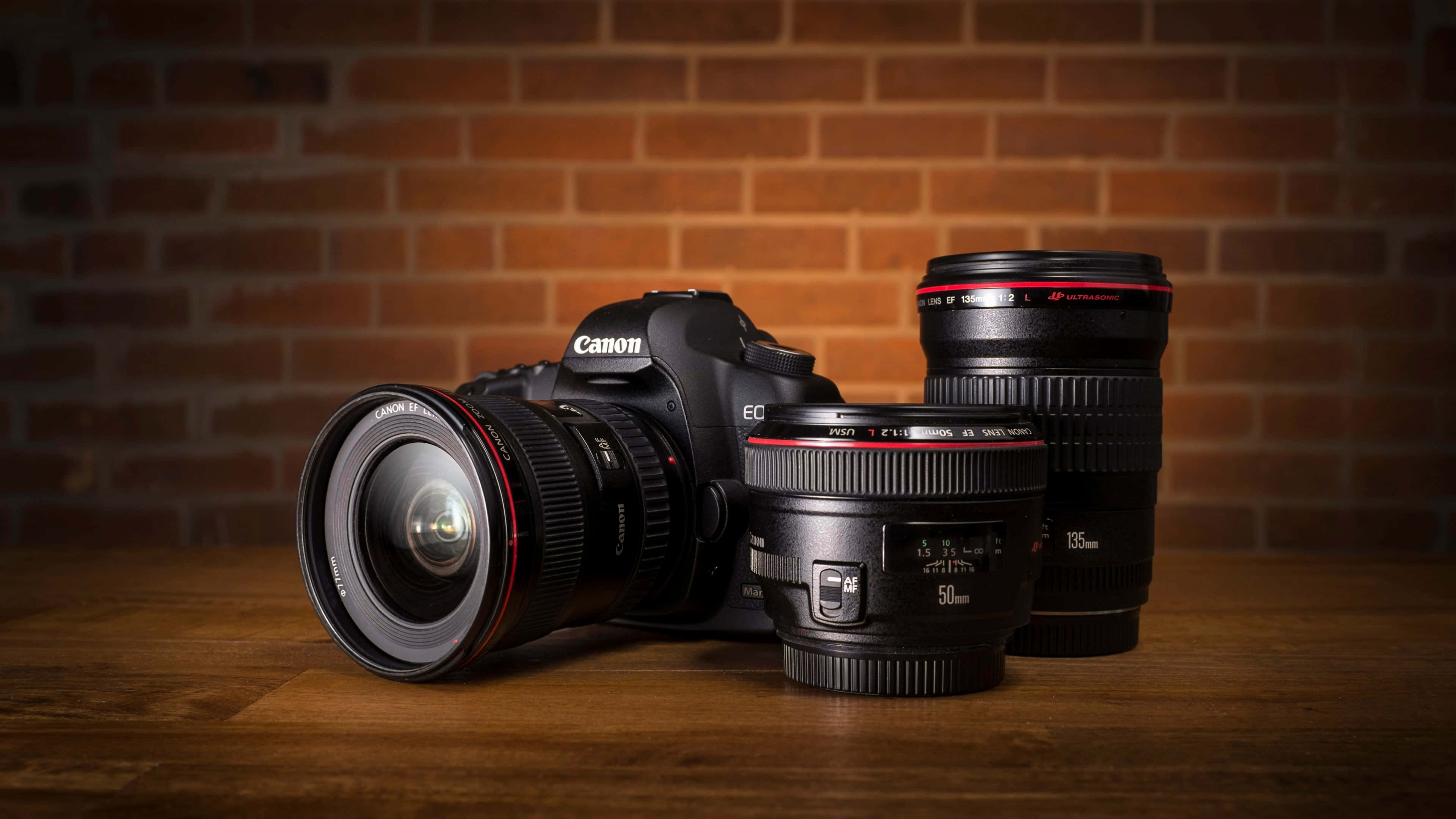 極高畫質 桌布 壁紙 — 一台 Canon DSLR 相機與三個鏡頭放置在木質表面上，背景是溫暖的磚牆。