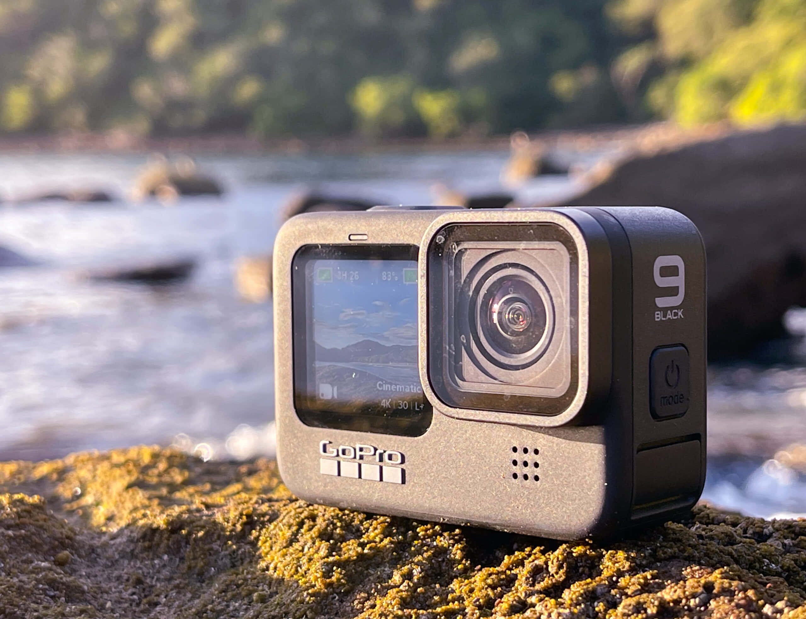 gopro 桌布 壁紙 — 一台 GoPro 9 Black 運動攝影機放在長滿青苔的石頭上，旁邊是一條流動的河流，背景是模糊的綠色森林。