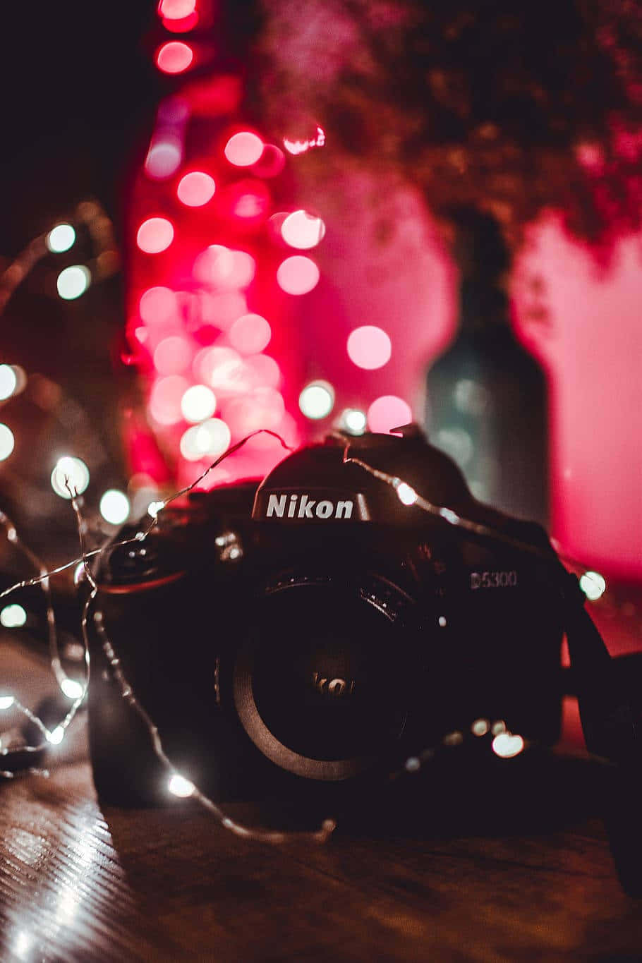 Dslr Camera 桌布 壁紙 — 一台 Nikon D5300 相機放置在木質表面上，夜晚被發光的紅色和白色仙女燈包圍。