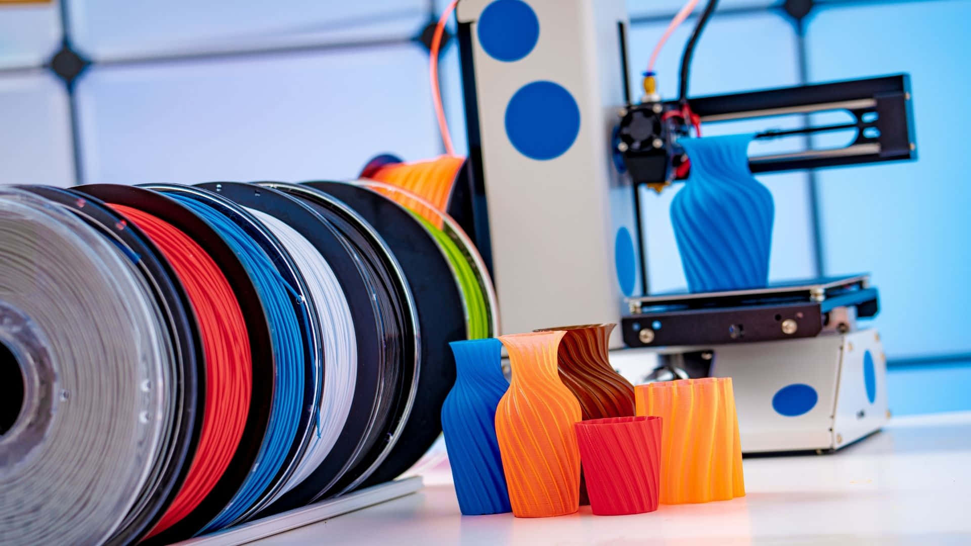 3d Printer 桌布 壁紙 — 一台3D列印機正在擠出藍色線材，而色彩繽紛的線軸和列印出來的花瓶則放在乾淨的白色工作檯面上。