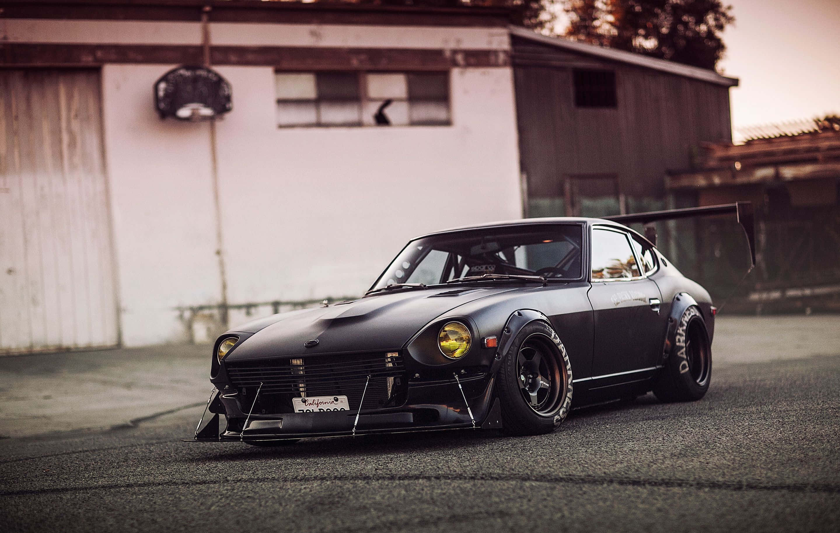Datsun 桌布 壁紙 — 一台啞光黑色改裝Datsun 240Z，配備寬輪胎和後尾翼，在黃昏時停放在風化車庫前。